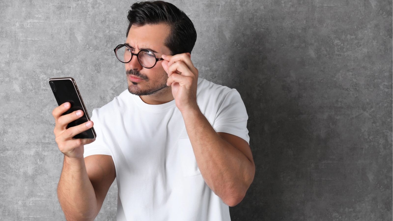 Man in a white t-shirt squints at his smartphone while adjusting his glasses, standing before a gray textured background.