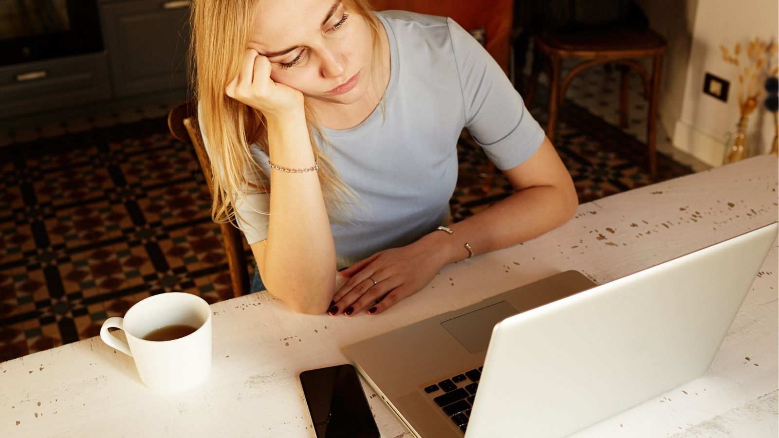 A woman rests her head on her hand while looking at a laptop, with a coffee and smartphone beside her on the table.