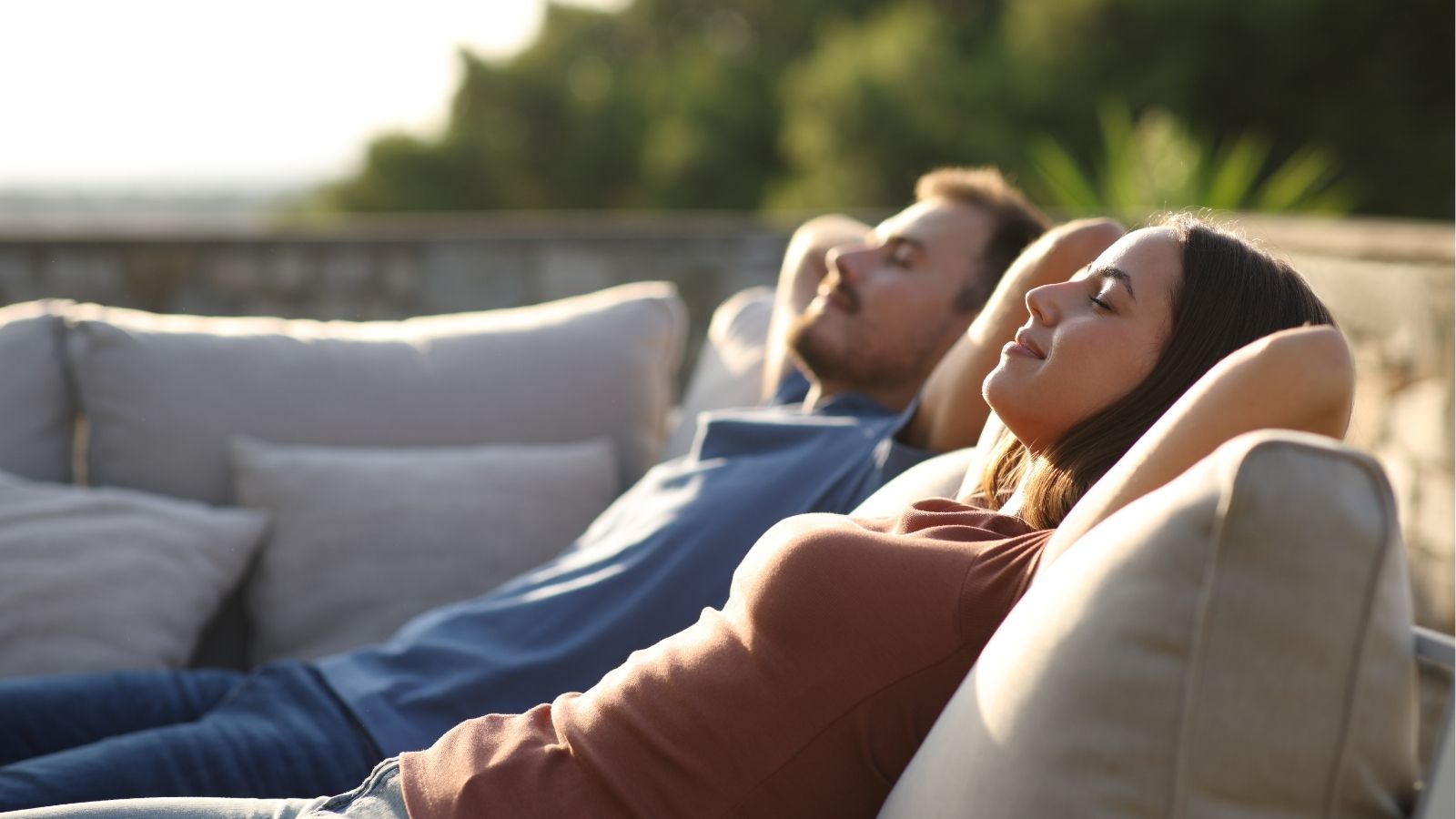 Two people recline on an outdoor sofa, eyes closed and hands behind heads, soaking up the sunlight.