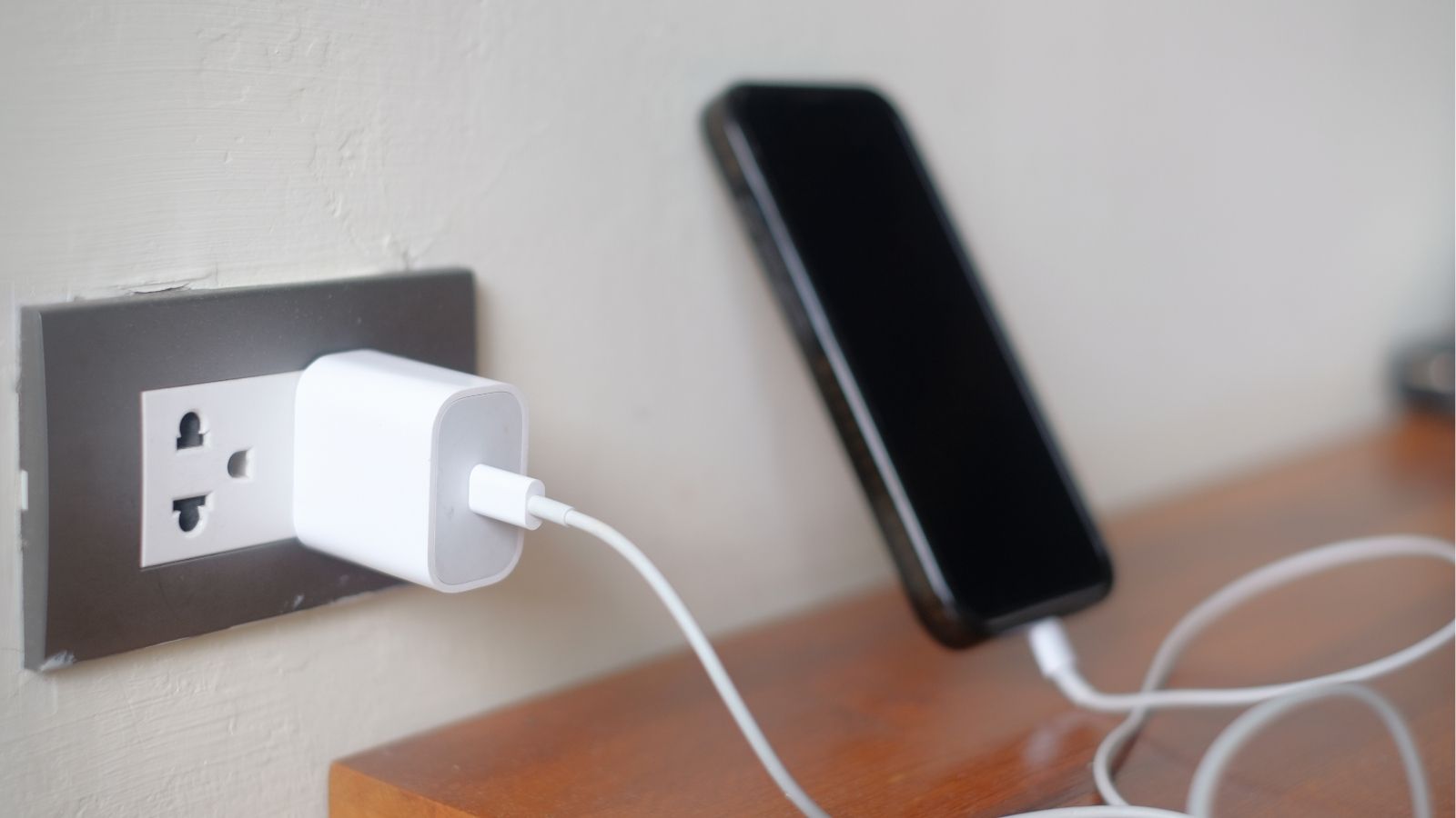 A smartphone charges on a wooden surface, plugged into a wall outlet with a cable and power adapter.