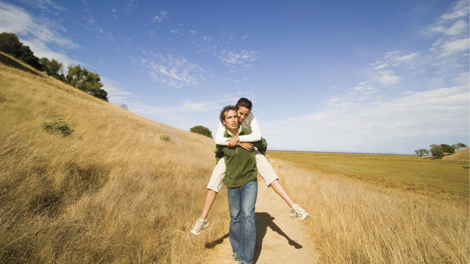 One person gives another a piggyback ride on a dirt path through a dry, grassy field under a blue sky with scattered clouds.