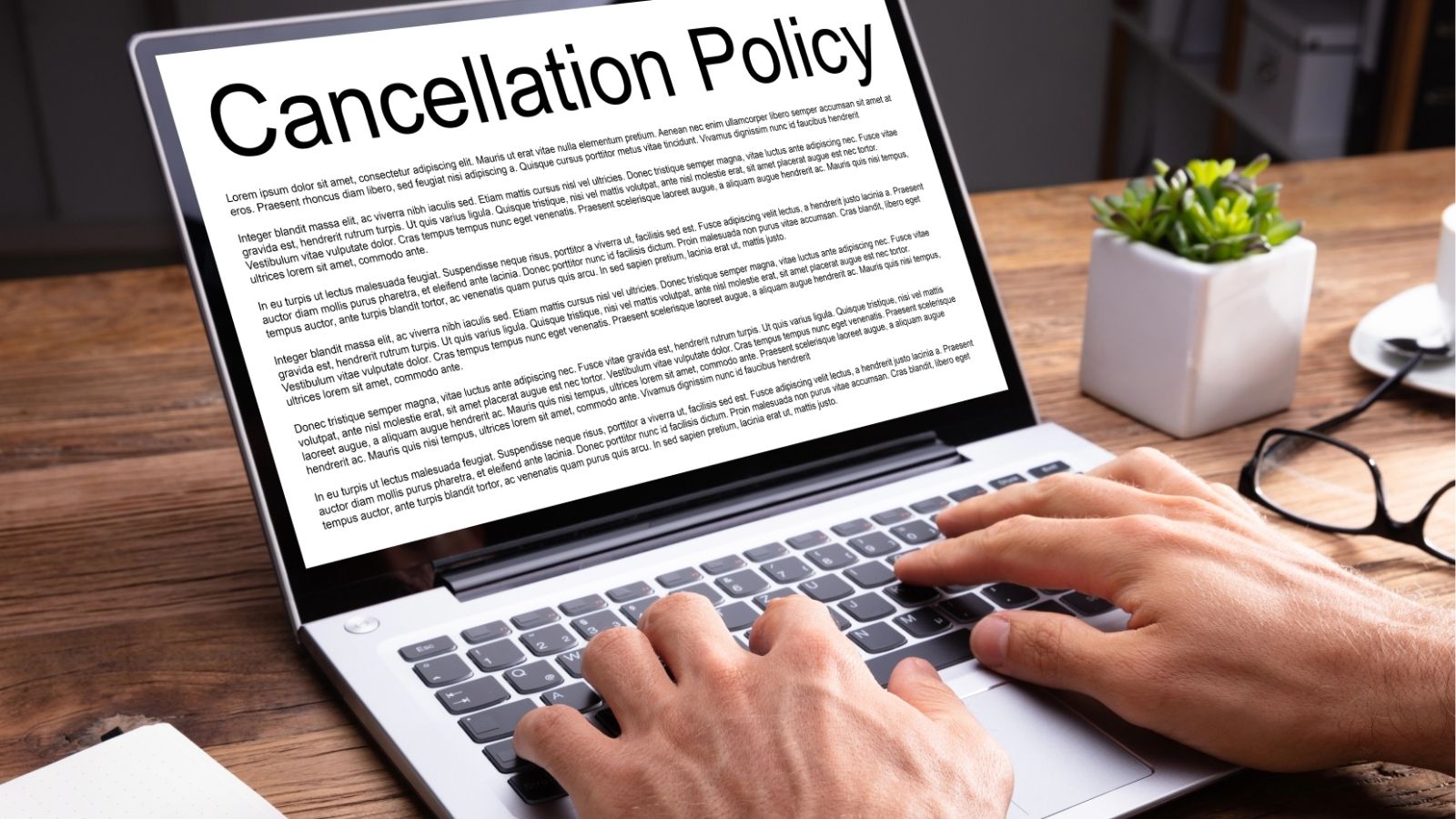 A person types on a laptop showing a "Cancellation Policy" document, with a plant and notebook also on the desk.