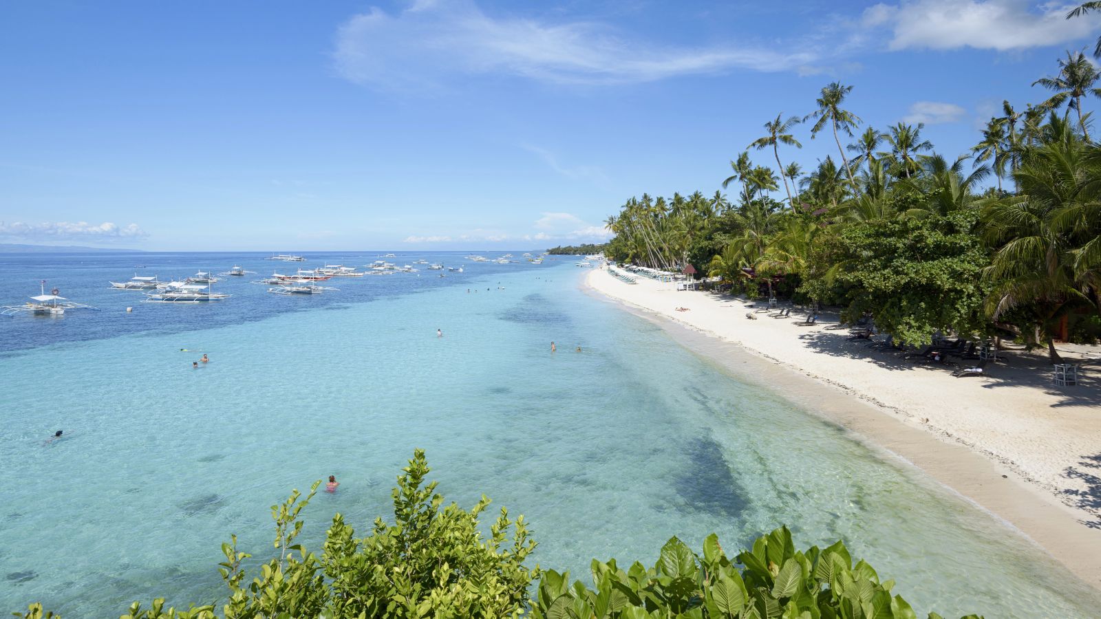 A sunny tropical beach with clear blue water, white sand, palm trees, and several boats anchored just offshore.