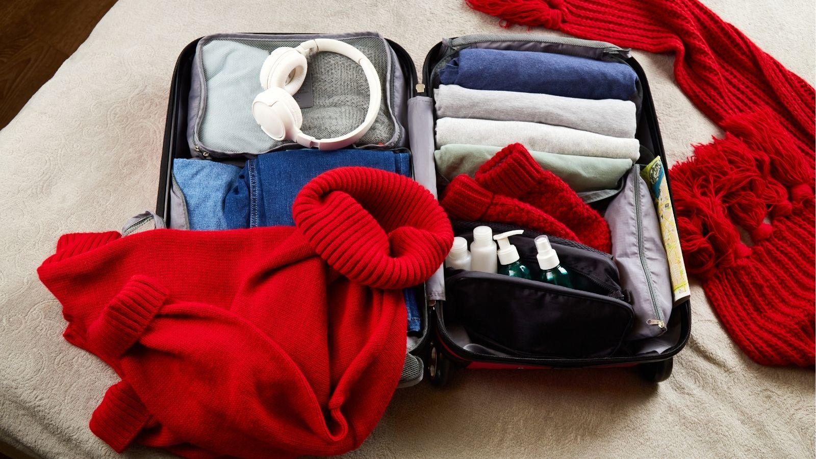 Open suitcase on a bed with folded clothes, headphones, toiletries, and a red sweater and scarf over the side.