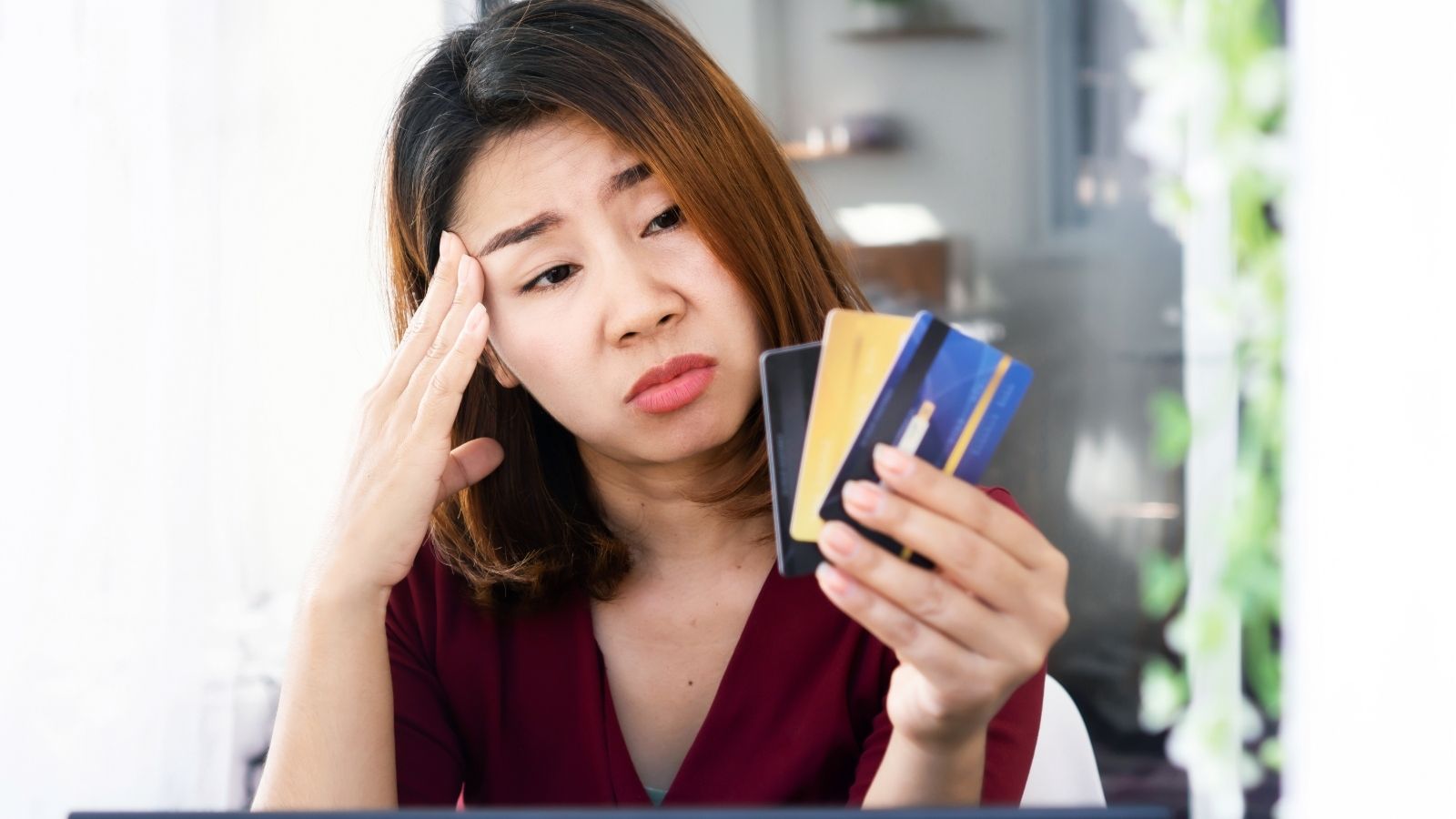A worried woman in a maroon shirt holds credit cards in one hand and touches her forehead with the other.