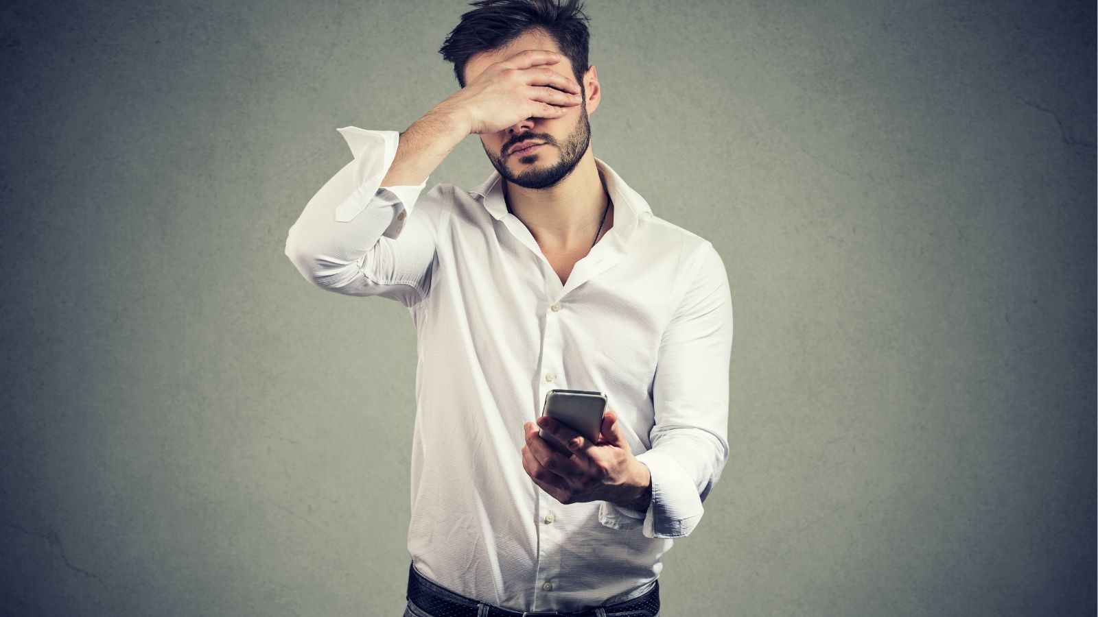 A man in a white shirt stands against a gray background, holding a smartphone and covering his face with one hand.