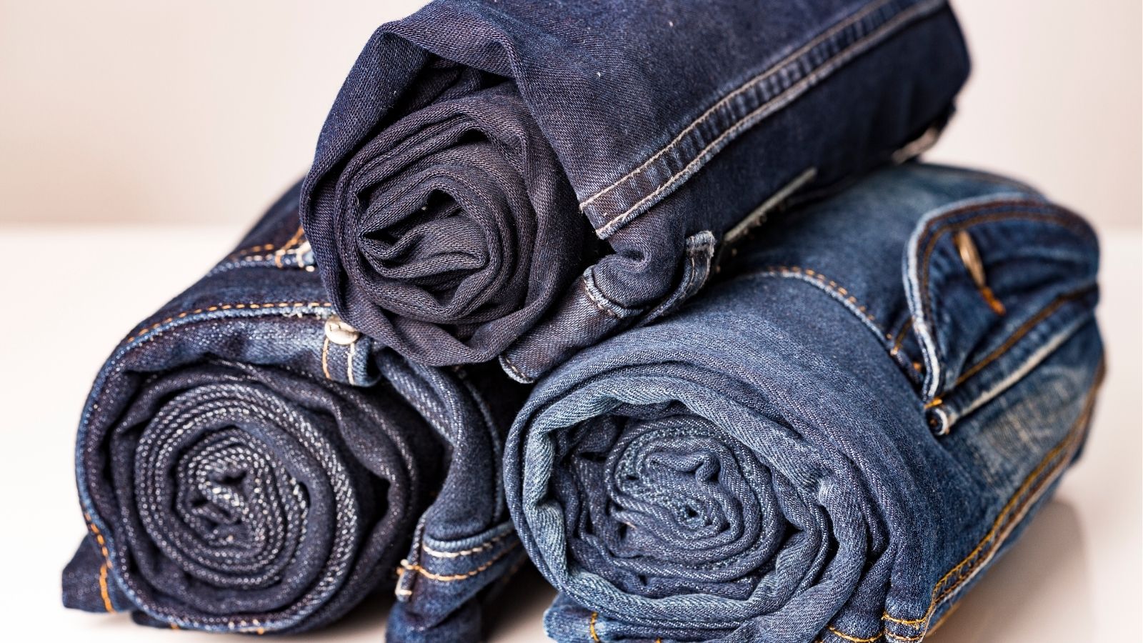 Three rolled-up blue jeans are stacked on a white surface.