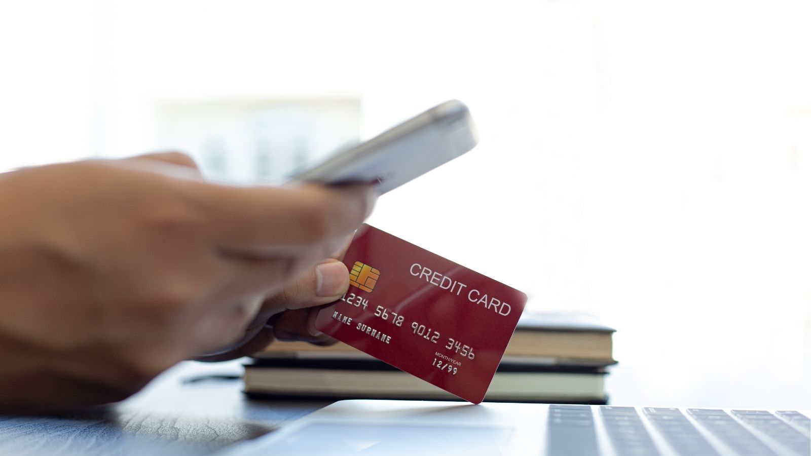 Person holds a credit card and smartphone near a laptop, with stacked books visible in the background.
