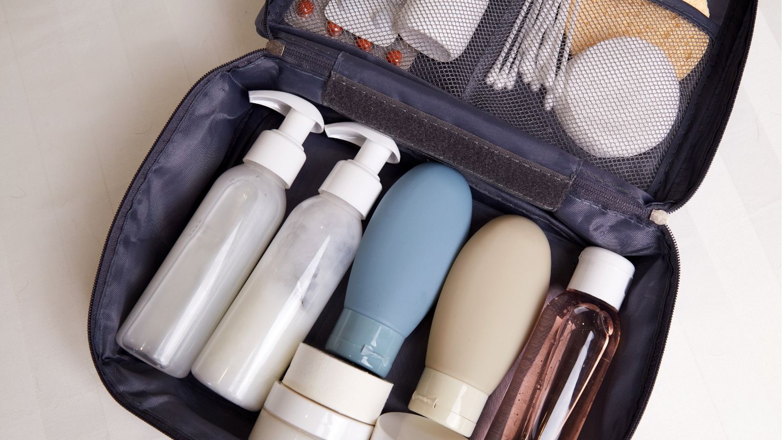 Open toiletry bag neatly holding pump bottles, a blue bottle, tubes, cotton swabs, pads, and small containers in compartments.