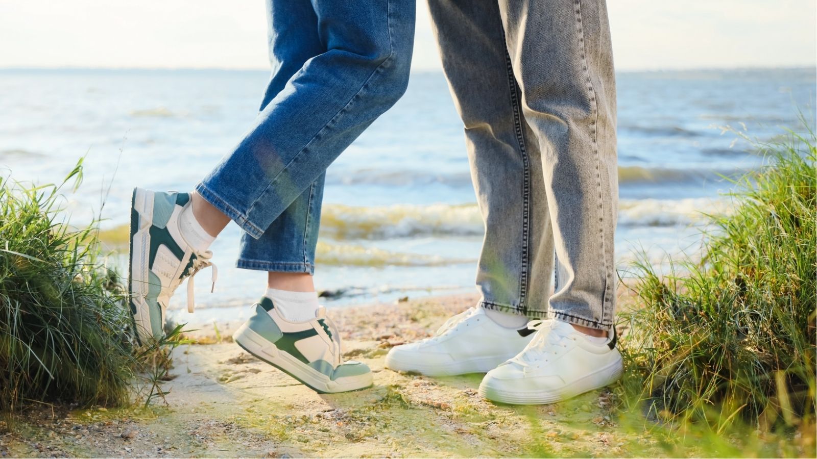 Two people in jeans and sneakers stand close on a grassy shoreline by water, one person on tiptoe.
