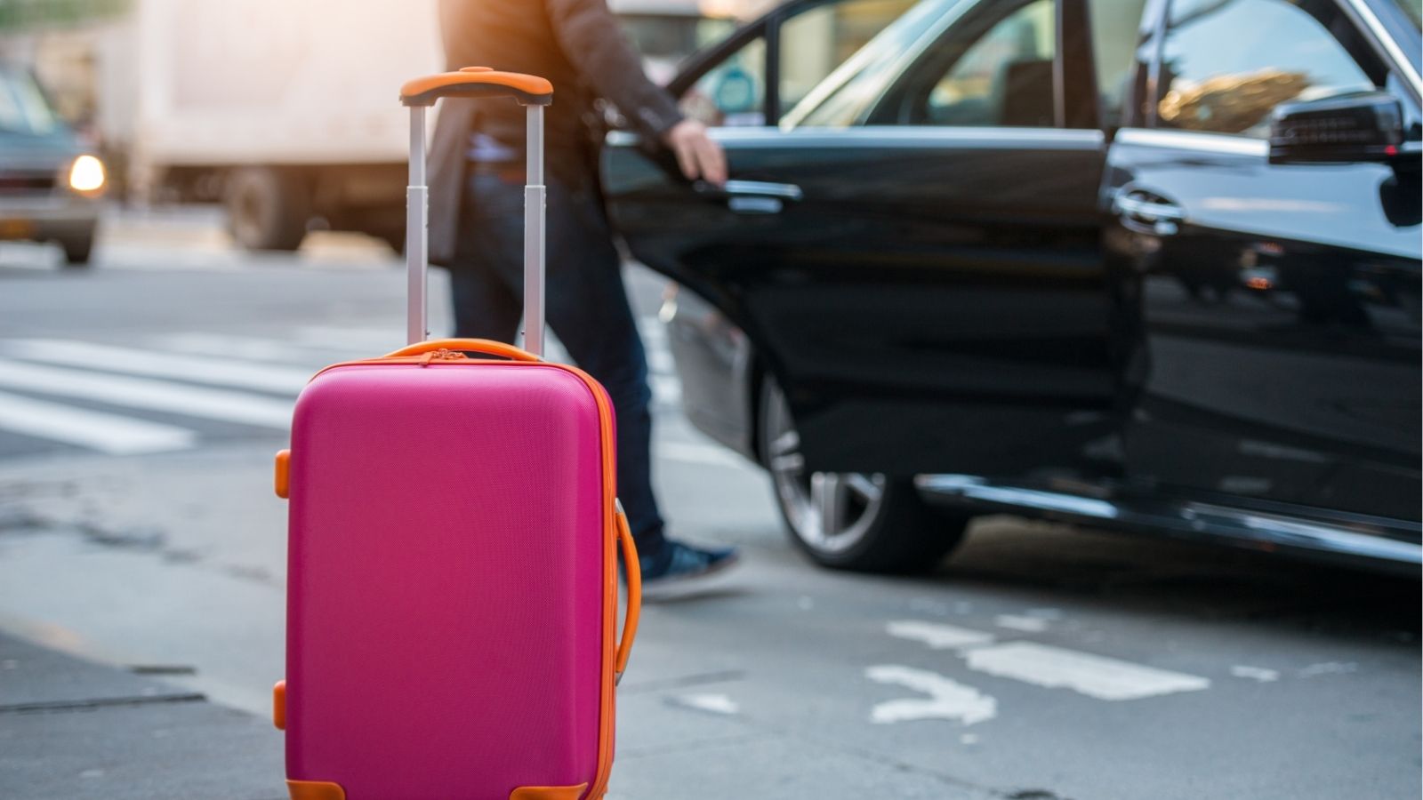 Bright pink suitcase with orange accents sits on a sidewalk as someone opens a black car door in the background.