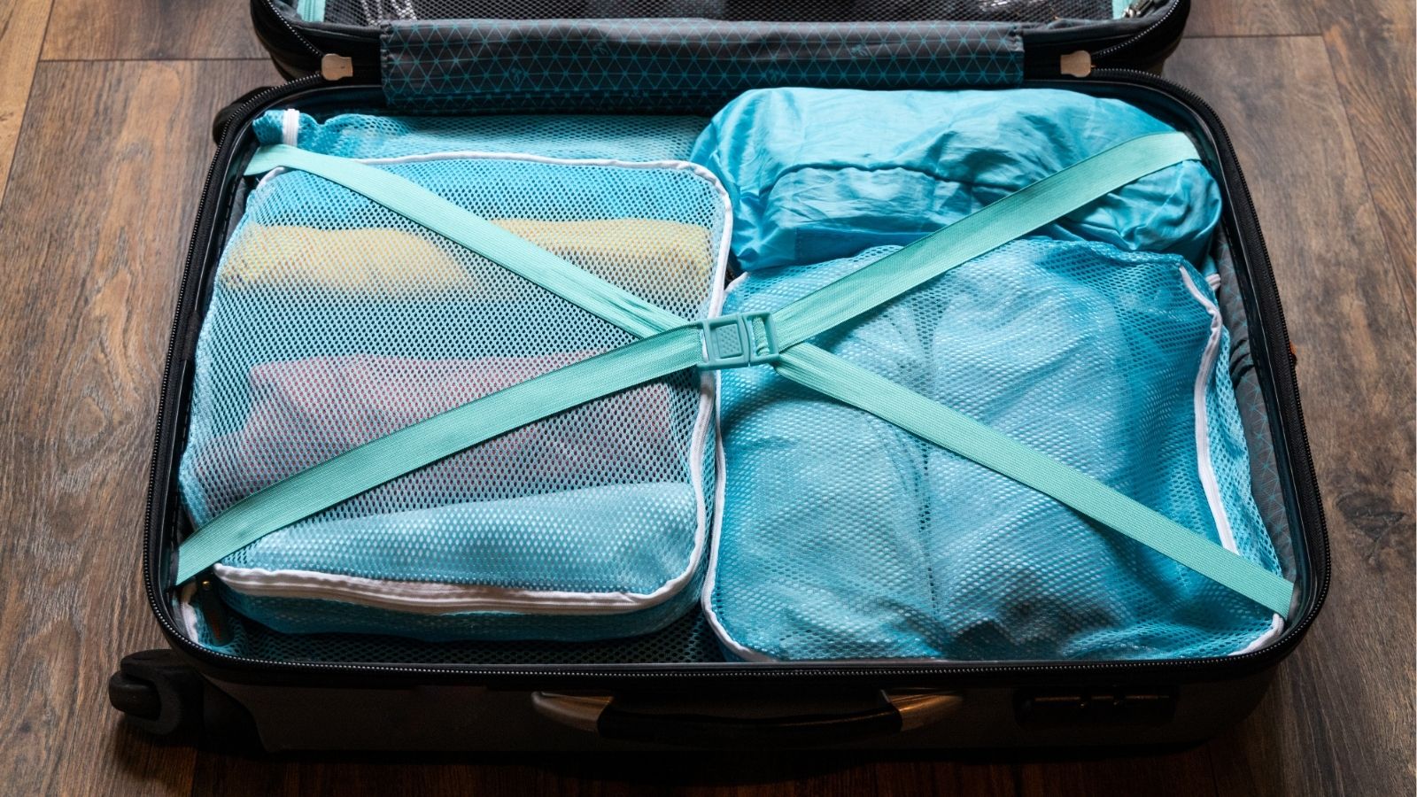An open suitcase with packing cubes and blue-strapped clothes sits on a wooden floor.