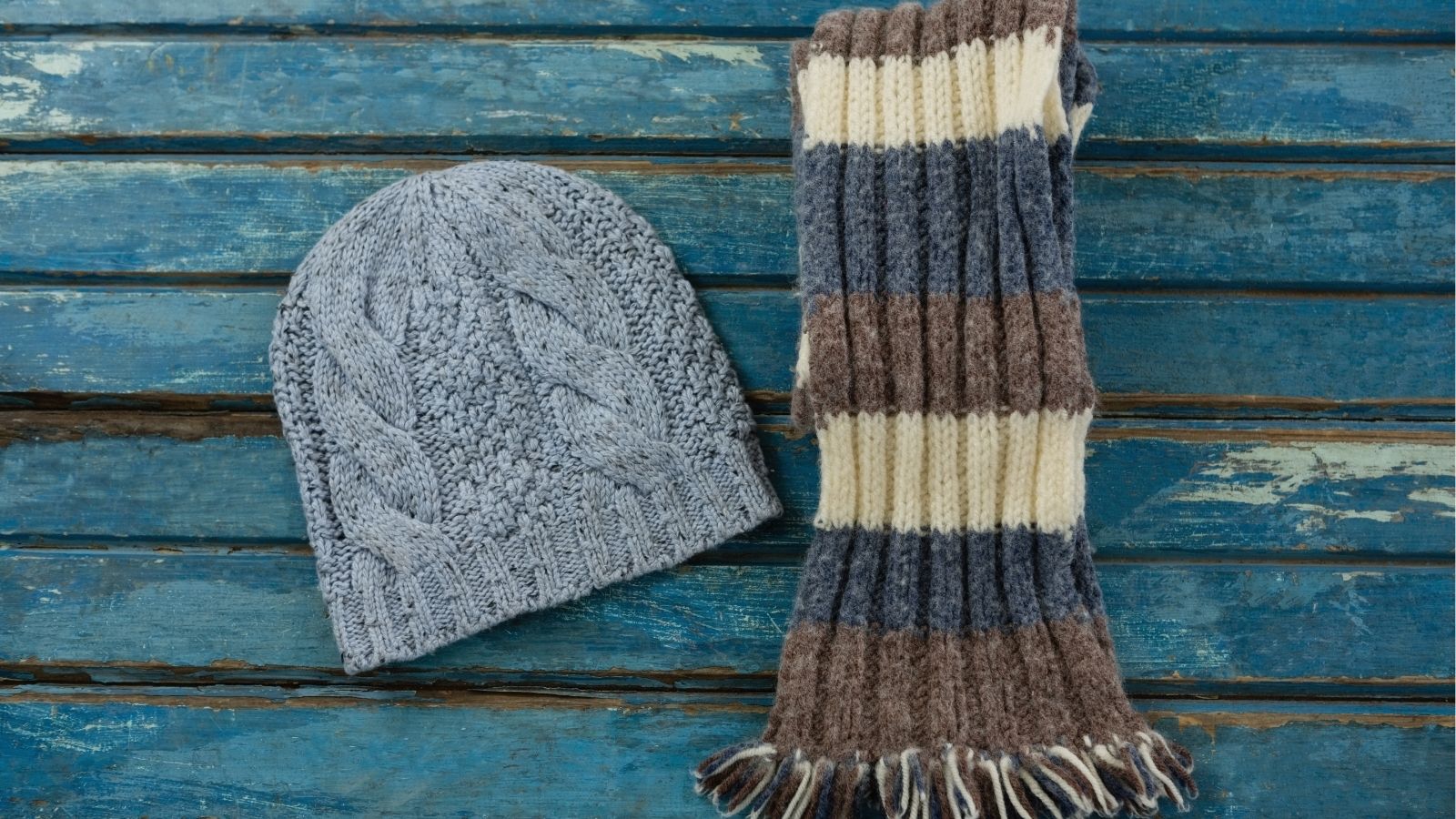 A gray knitted beanie and a gray, cream, and brown striped scarf rest on a weathered blue wooden surface.