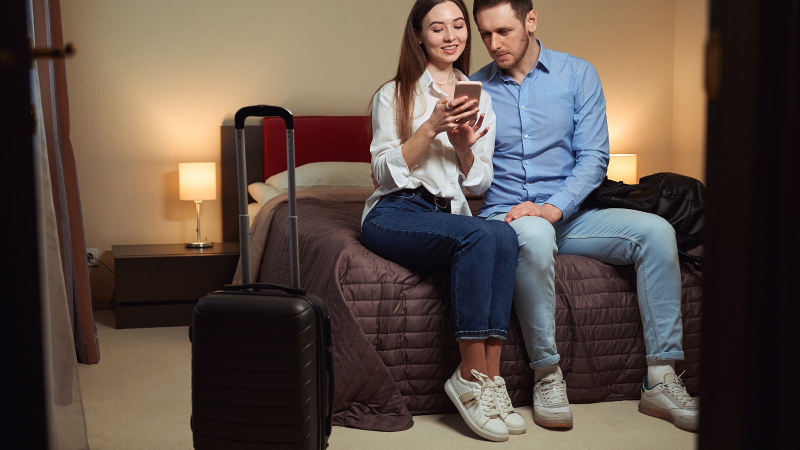 A photo of two couple sitting in the bed with a smile looking at woman's phone.