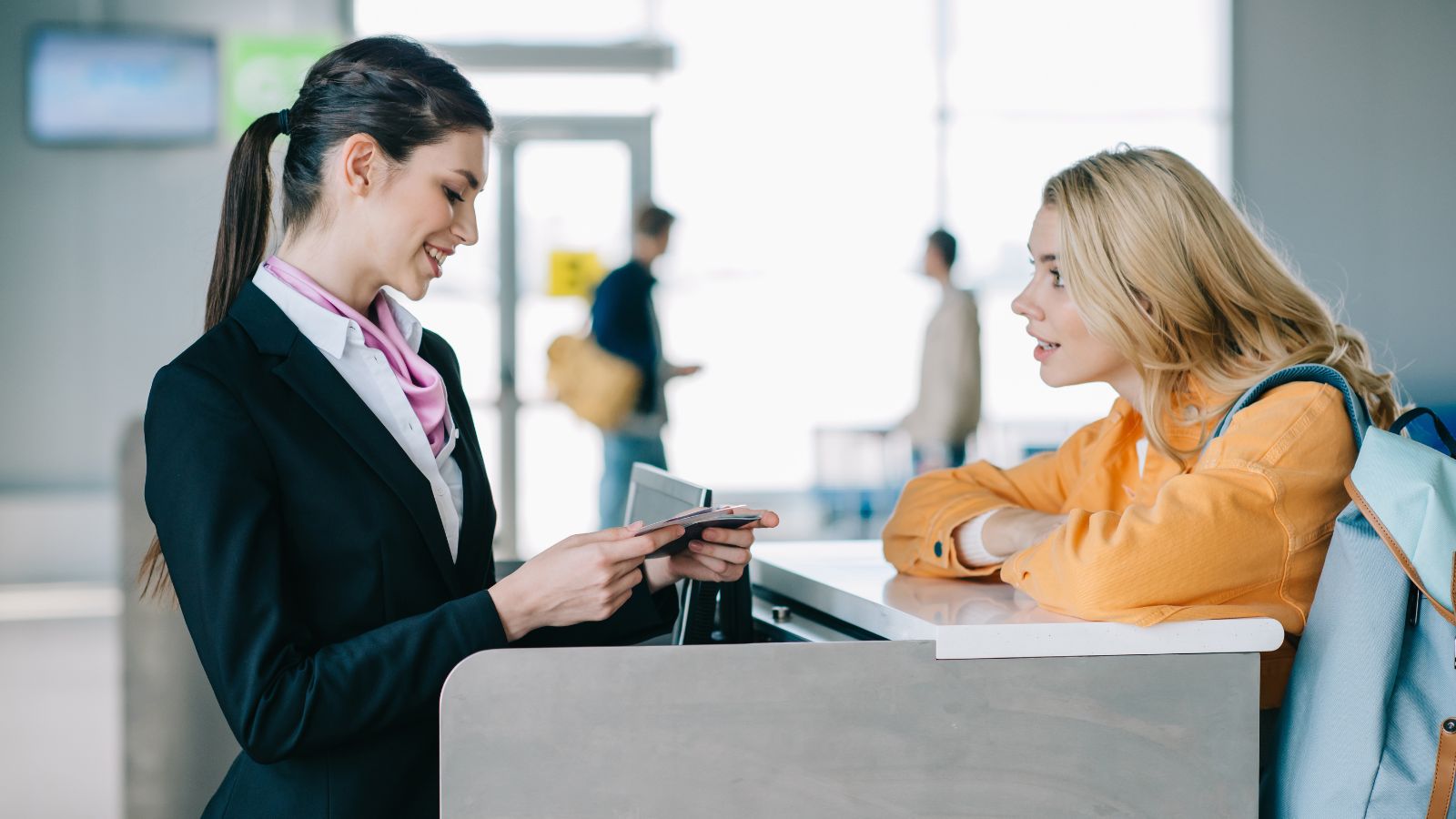 An airline staff member checks a passenger’s documents at the counter; the passenger leans with a backpack.
