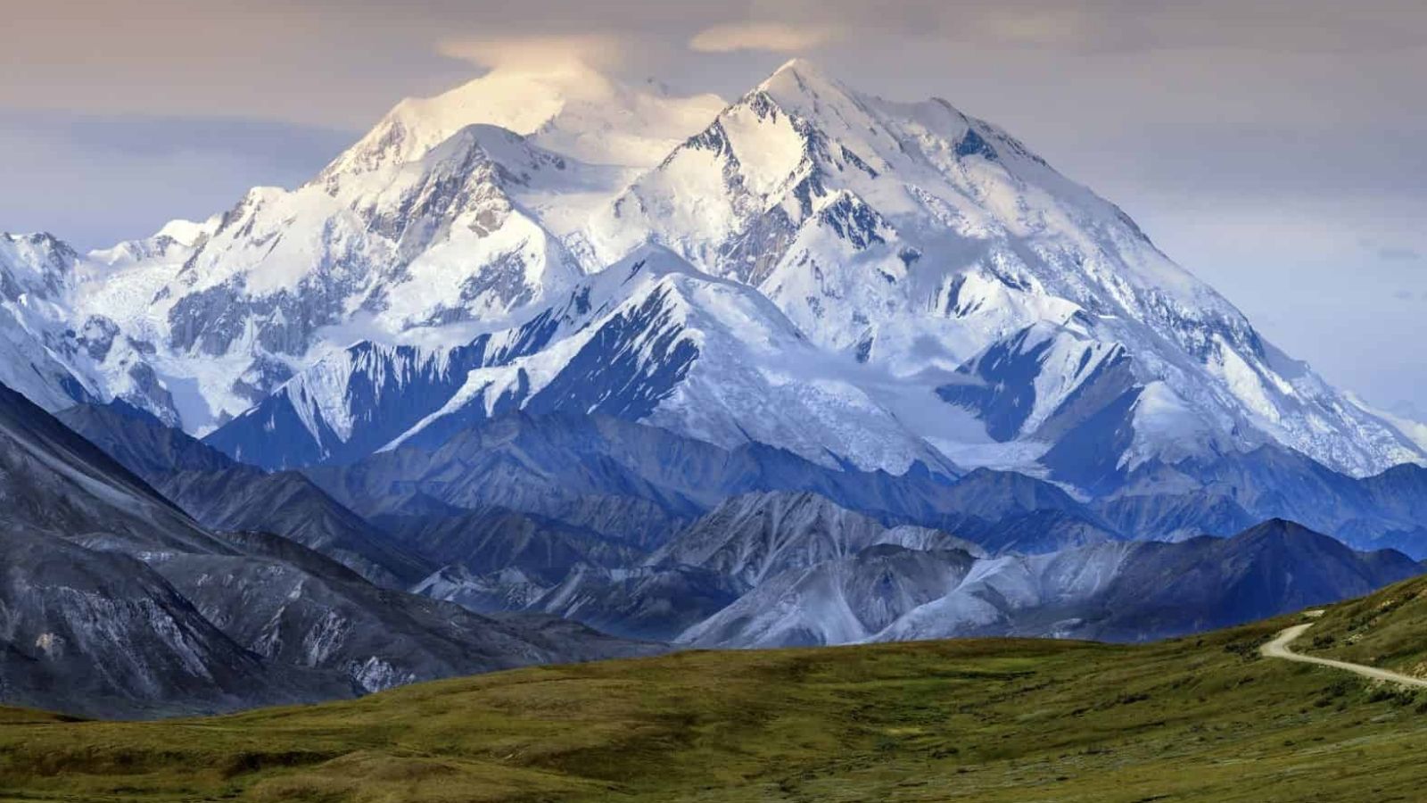 A snow-capped mountain towers over rocky slopes and grassy ground beneath a cloudy sky.