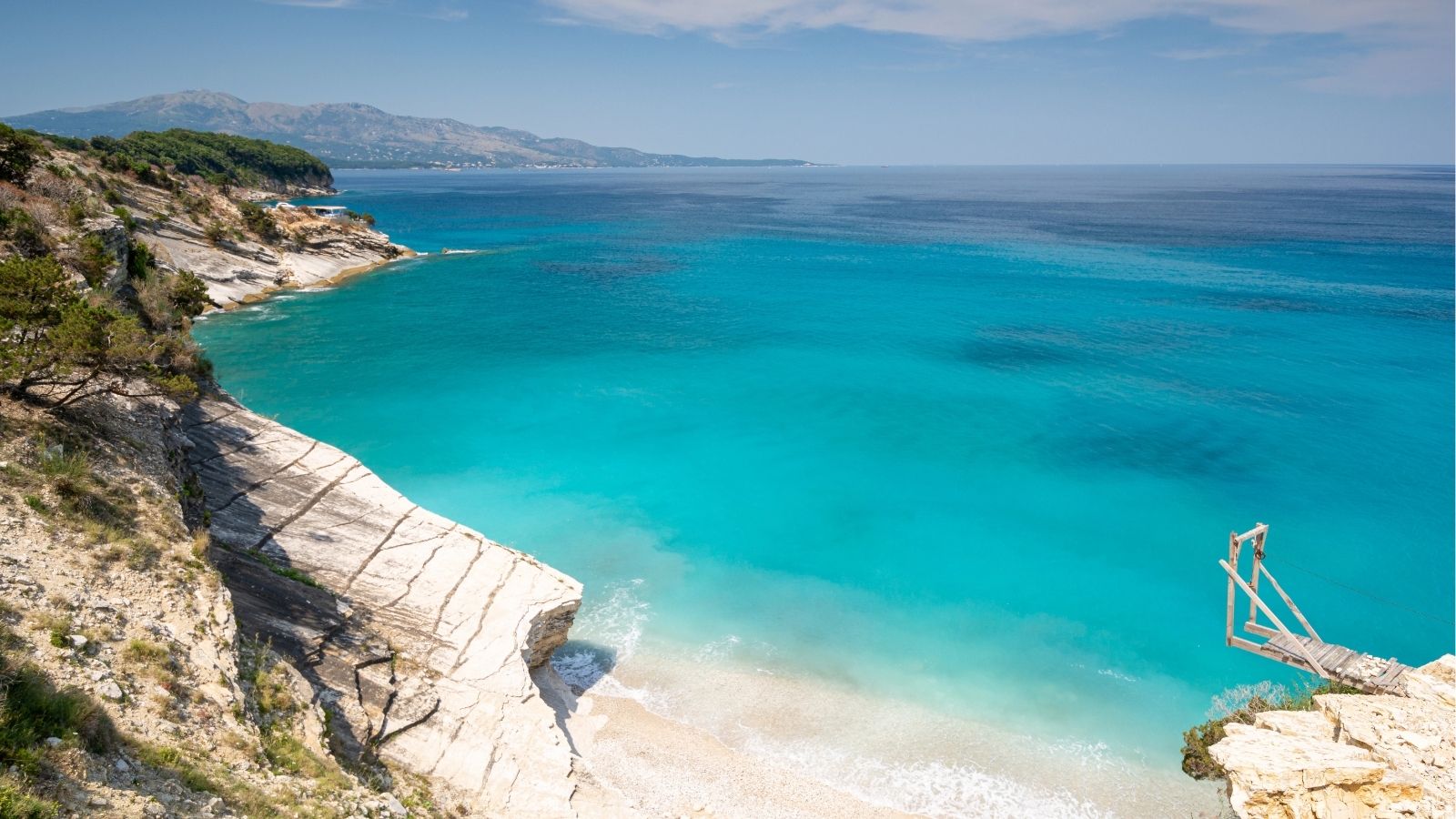 A rocky white coastline, turquoise water, sandy beach, and distant hills beneath a partly cloudy sky.