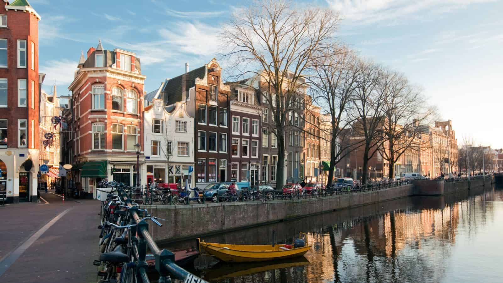 Amsterdam canal with bicycles, tall narrow buildings, and a yellow boat reflected in the water under a clear sky.