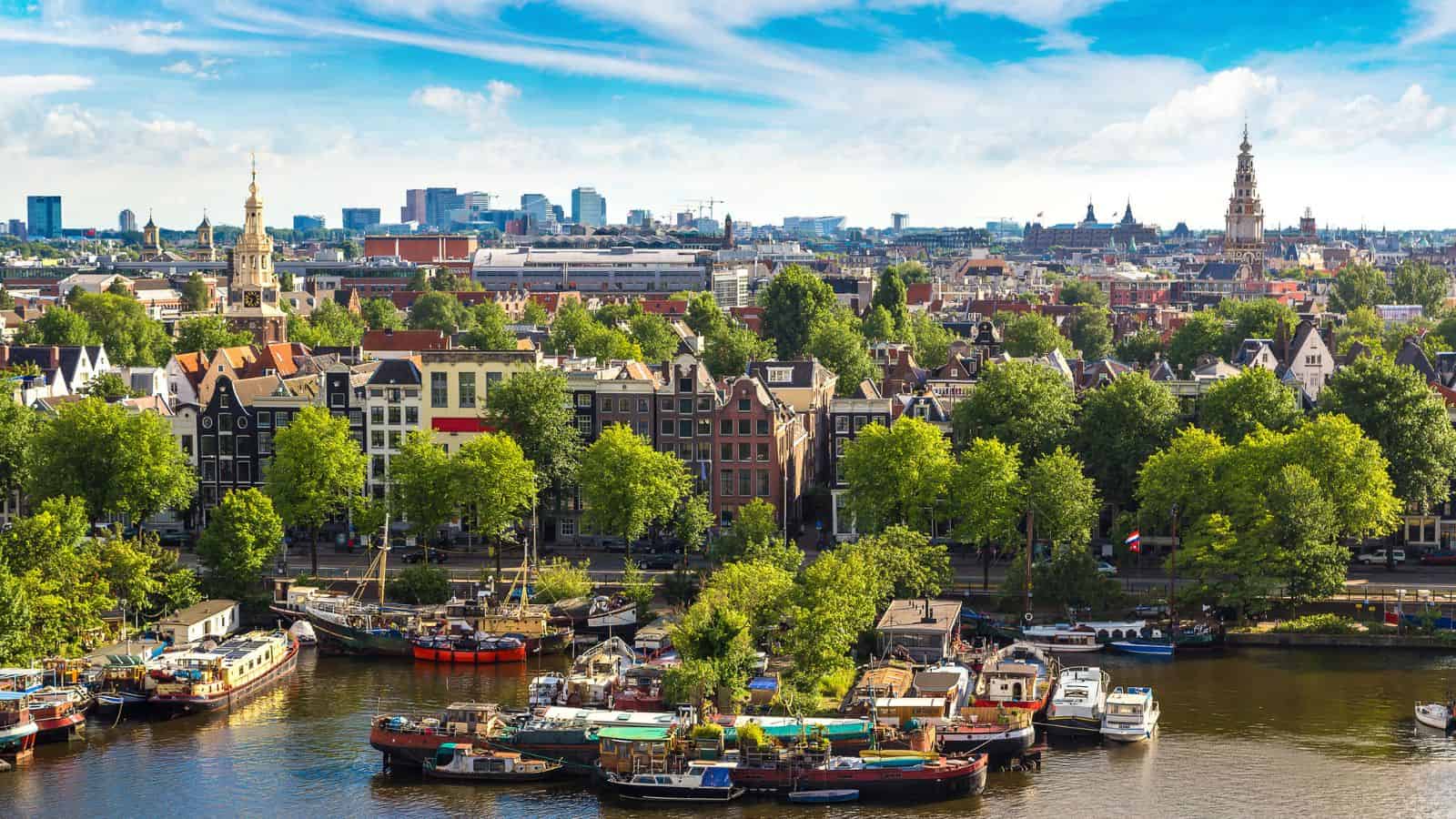 Boats and trees line a canal before traditional buildings beneath a partly cloudy sky in an urban cityscape.