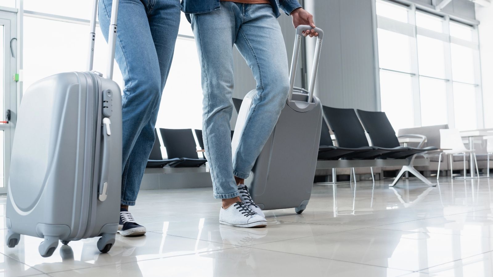 Two people in jeans and sneakers pull gray suitcases through an airport terminal with empty seats and large windows behind them.