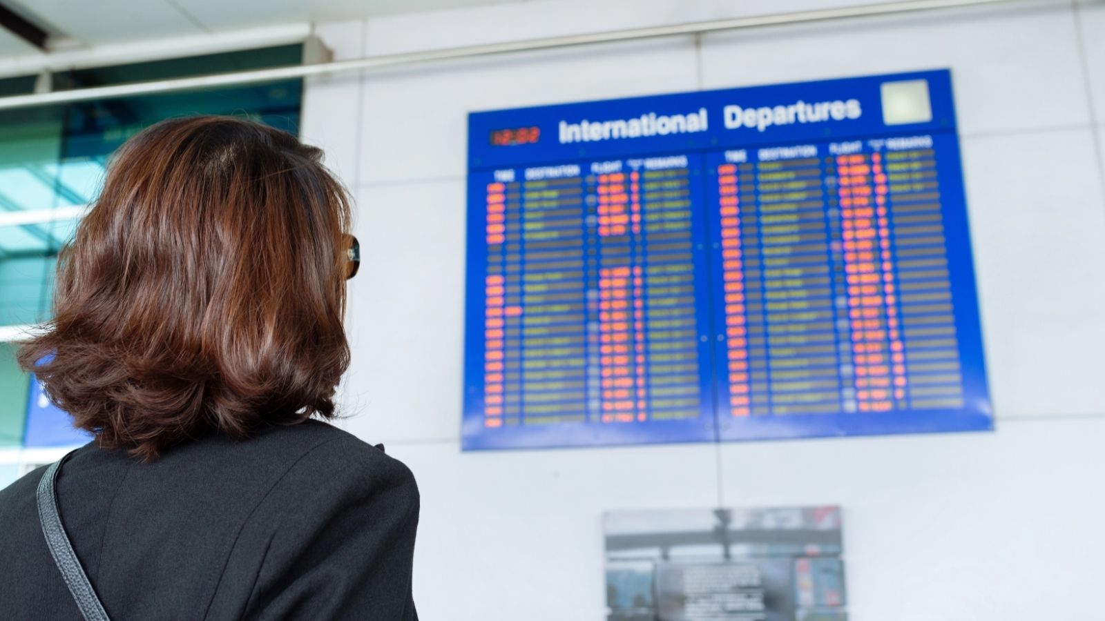 A person with shoulder-length hair views an international departures board at the airport.