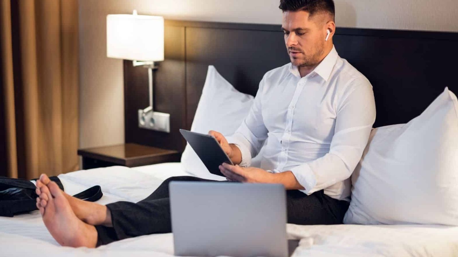 A man in business attire sits on a hotel bed using a tablet and wireless earbuds, with an open laptop in front of him.