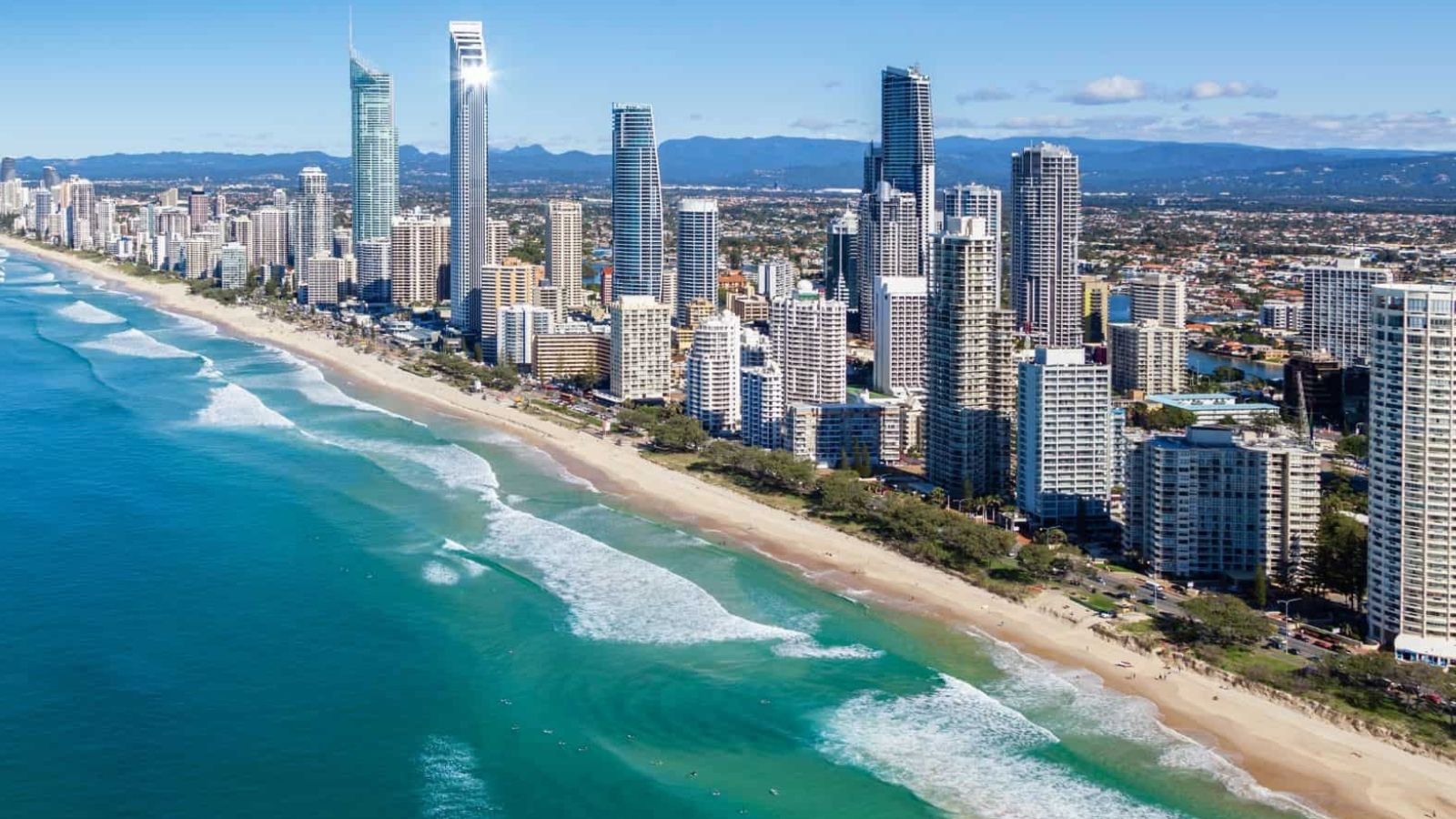 Aerial view of tall city buildings by a sandy beach and turquoise ocean beneath a clear blue sky.