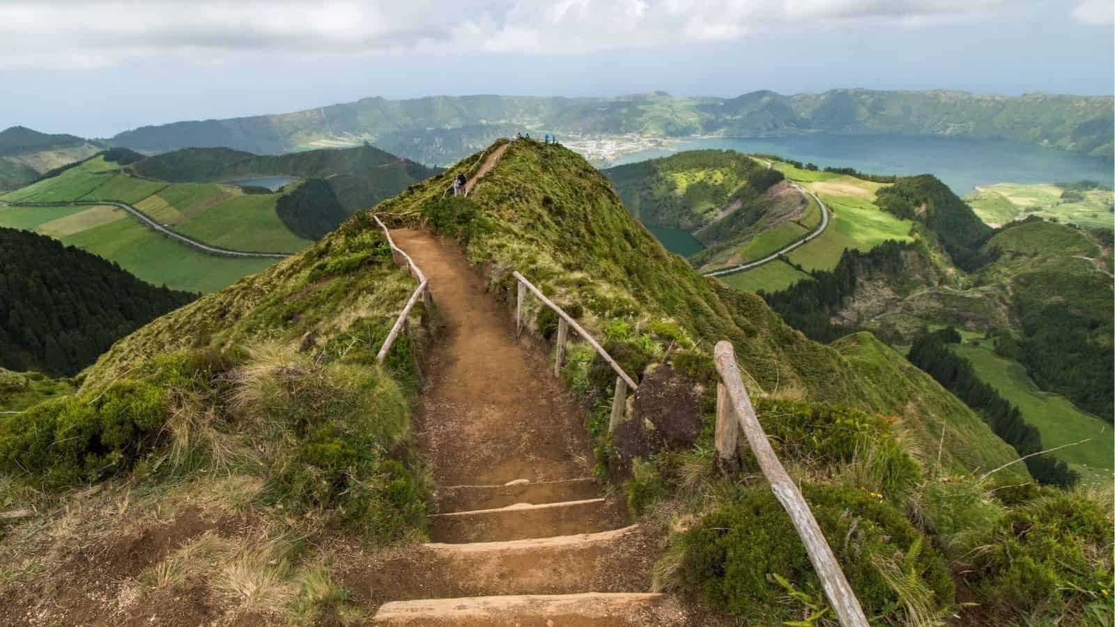 A dirt path with wooden railings ascends a ridge above green hills, a crater lake, and distant mountains beneath clouds.