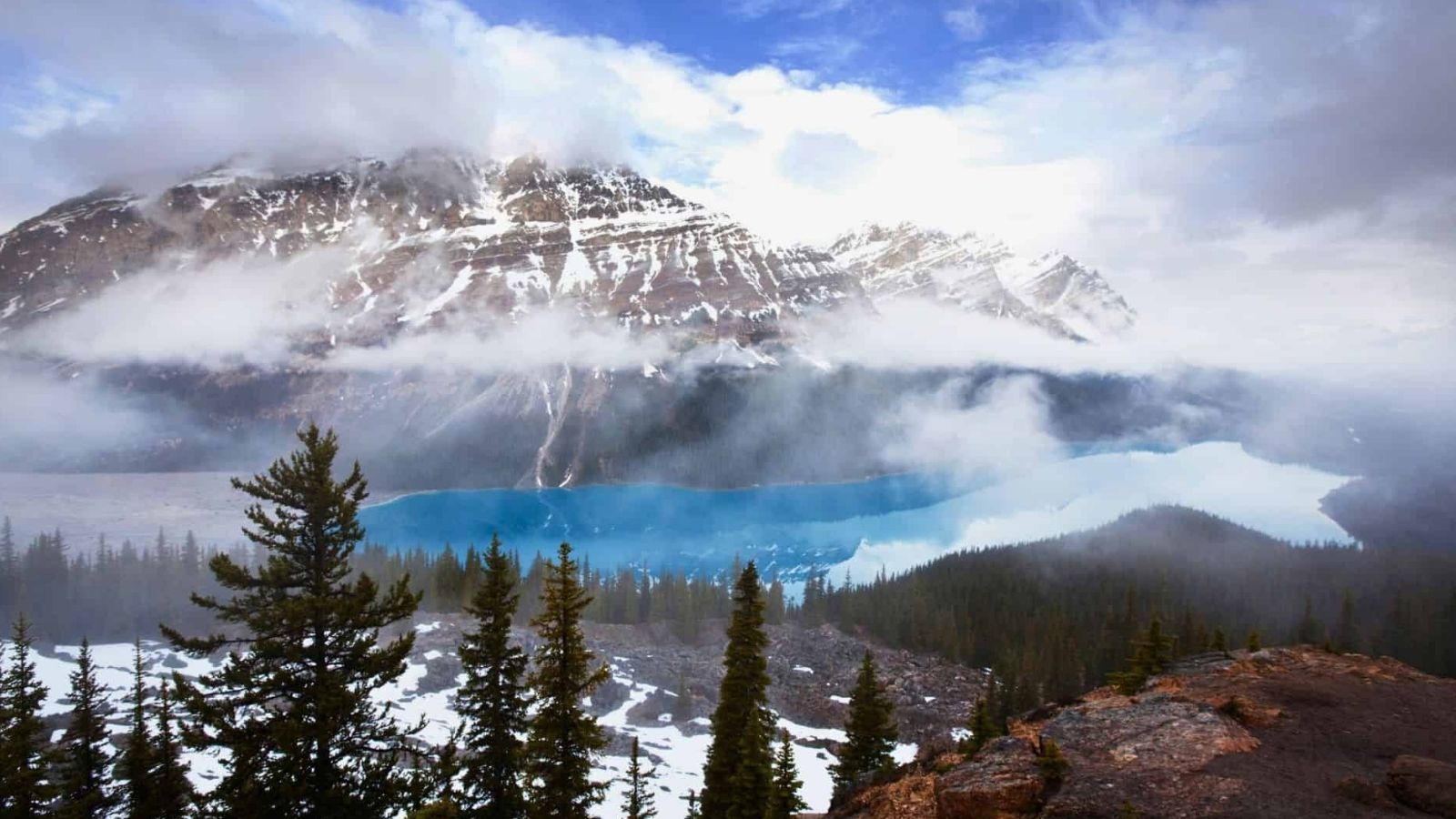 Snowy mountains shrouded in clouds overlook a blue lake, with evergreen trees in the foreground.