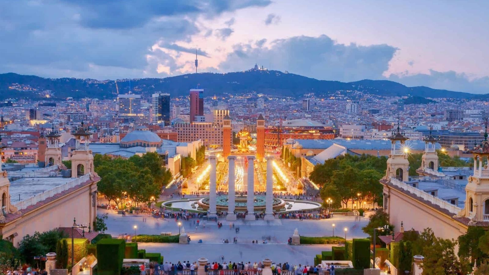 Barcelona at dusk with the lit Magic Fountain of Montjuïc, surrounding city buildings, and hills in the distance.