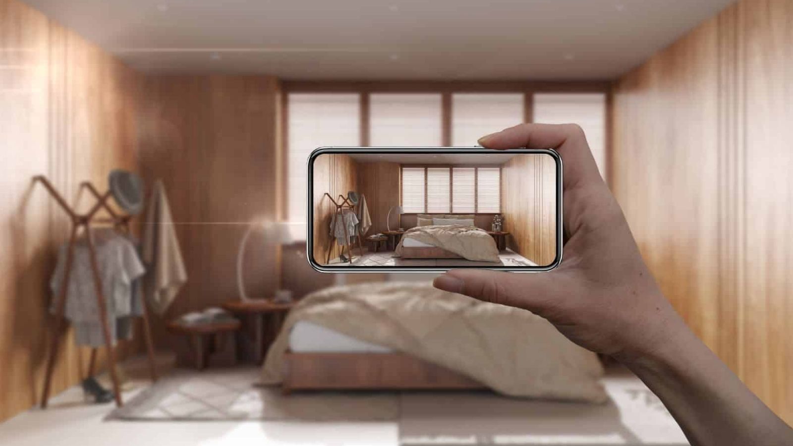 A hand holds a smartphone to photograph a modern wooden bedroom with an unmade bed and natural light.