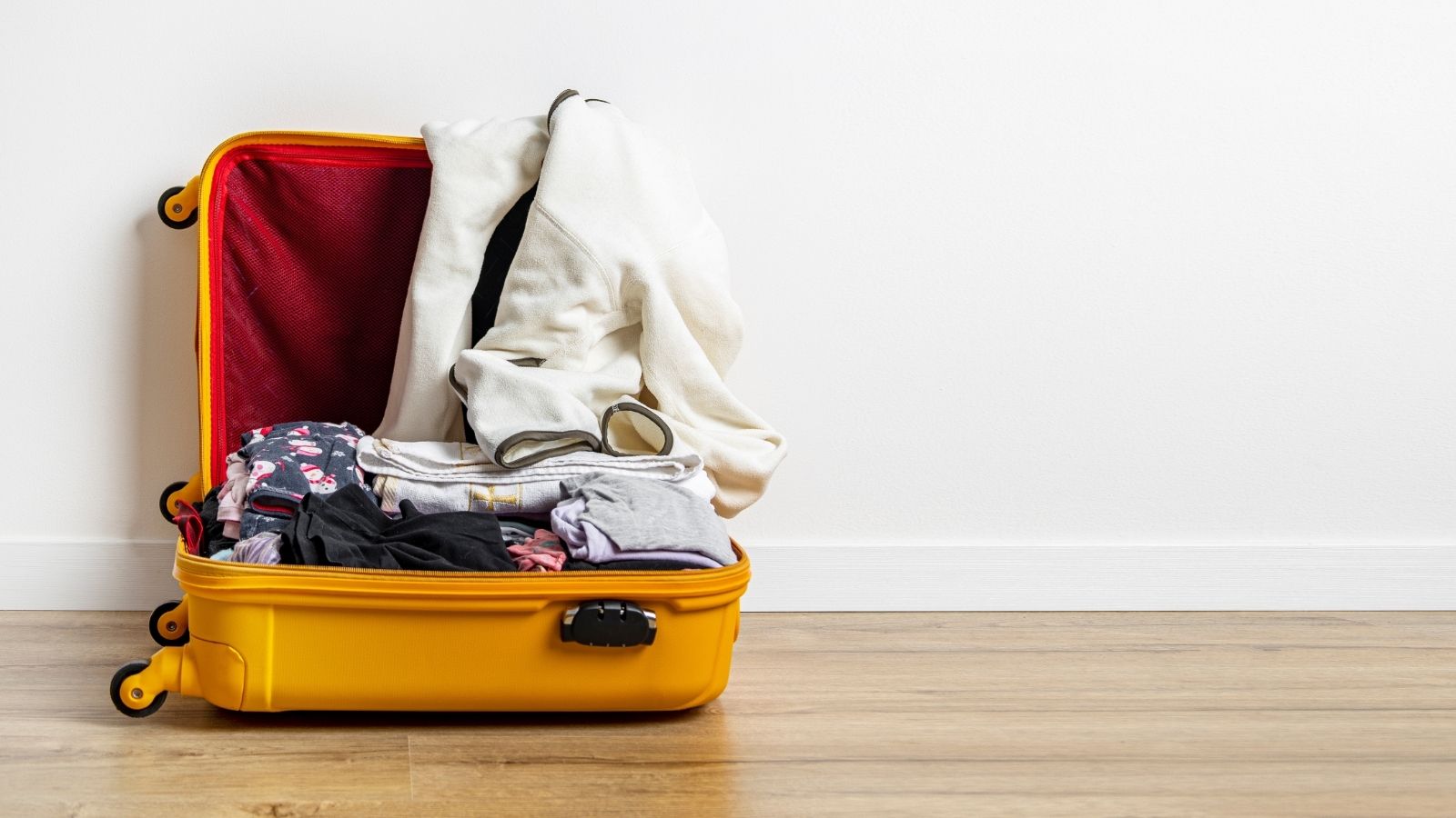 An open yellow suitcase containing clothes and sunglasses sits on a wooden floor by a white wall.