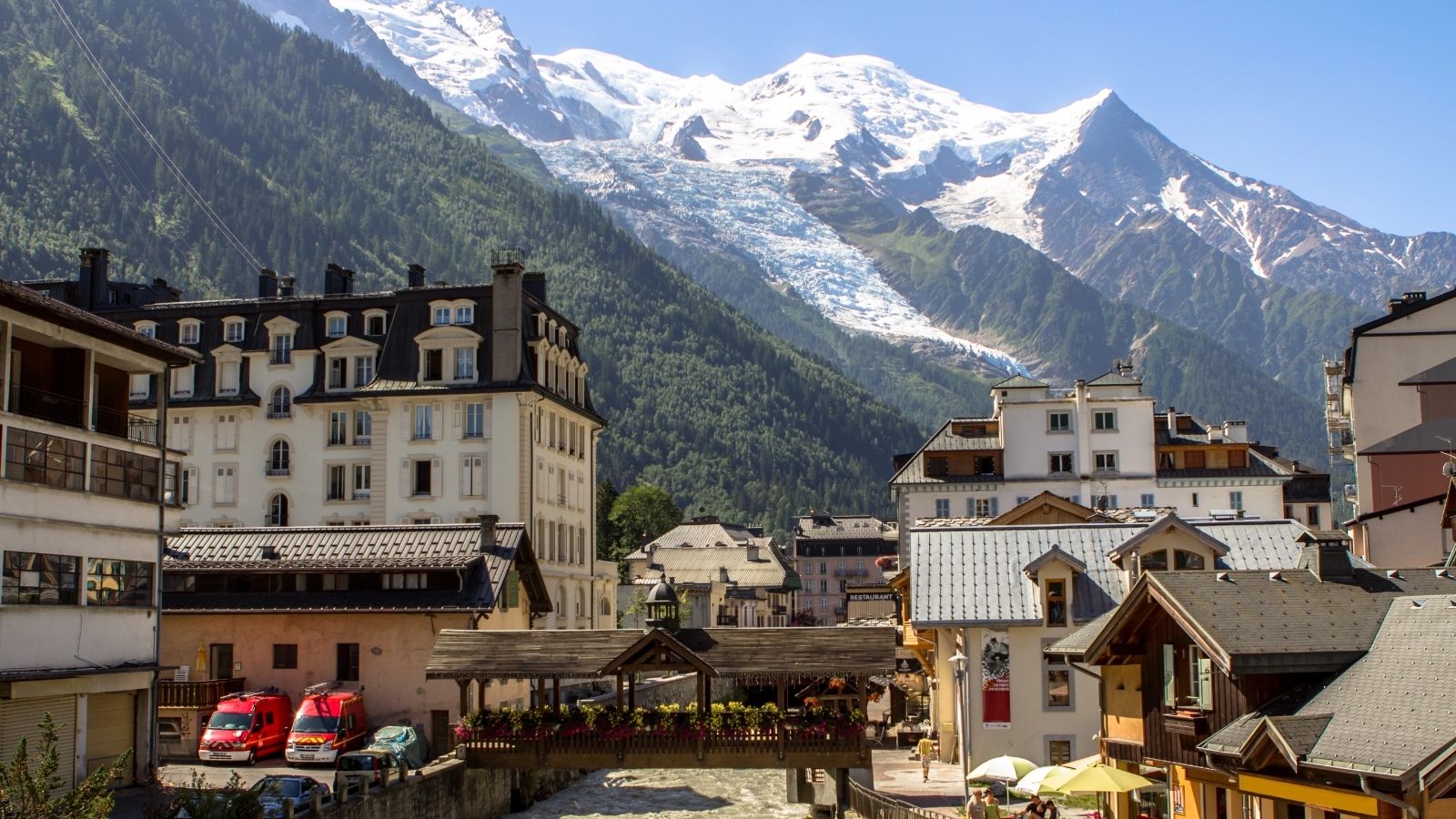 A mountain town’s buildings sit by a river, backed by snowy peaks and green forests beneath a clear blue sky.