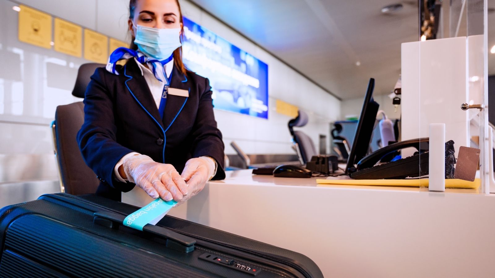 Masked, gloved airline staff attaches a baggage tag to a suitcase at the airport check-in counter.