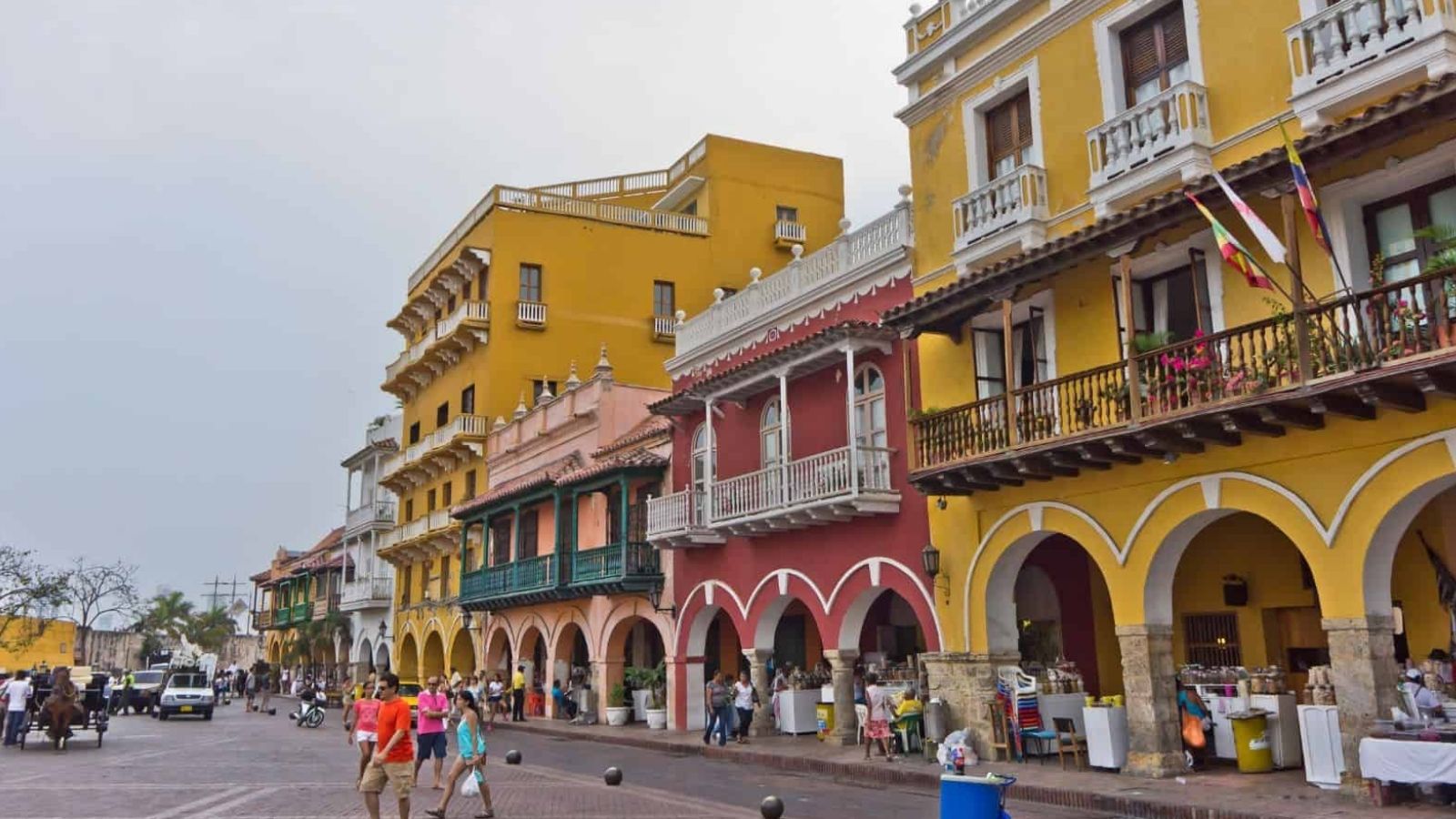 People walk past colorful colonial buildings with arches, balconies, and outdoor seating under a cloudy sky.