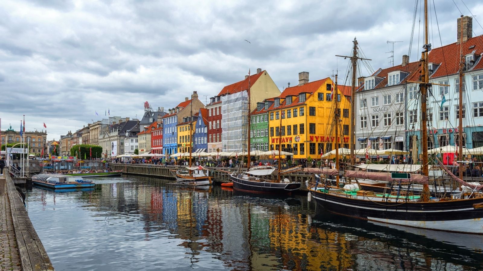 Colorful buildings, docked boats, and outdoor cafes border a canal beneath a cloudy sky on the city waterfront.