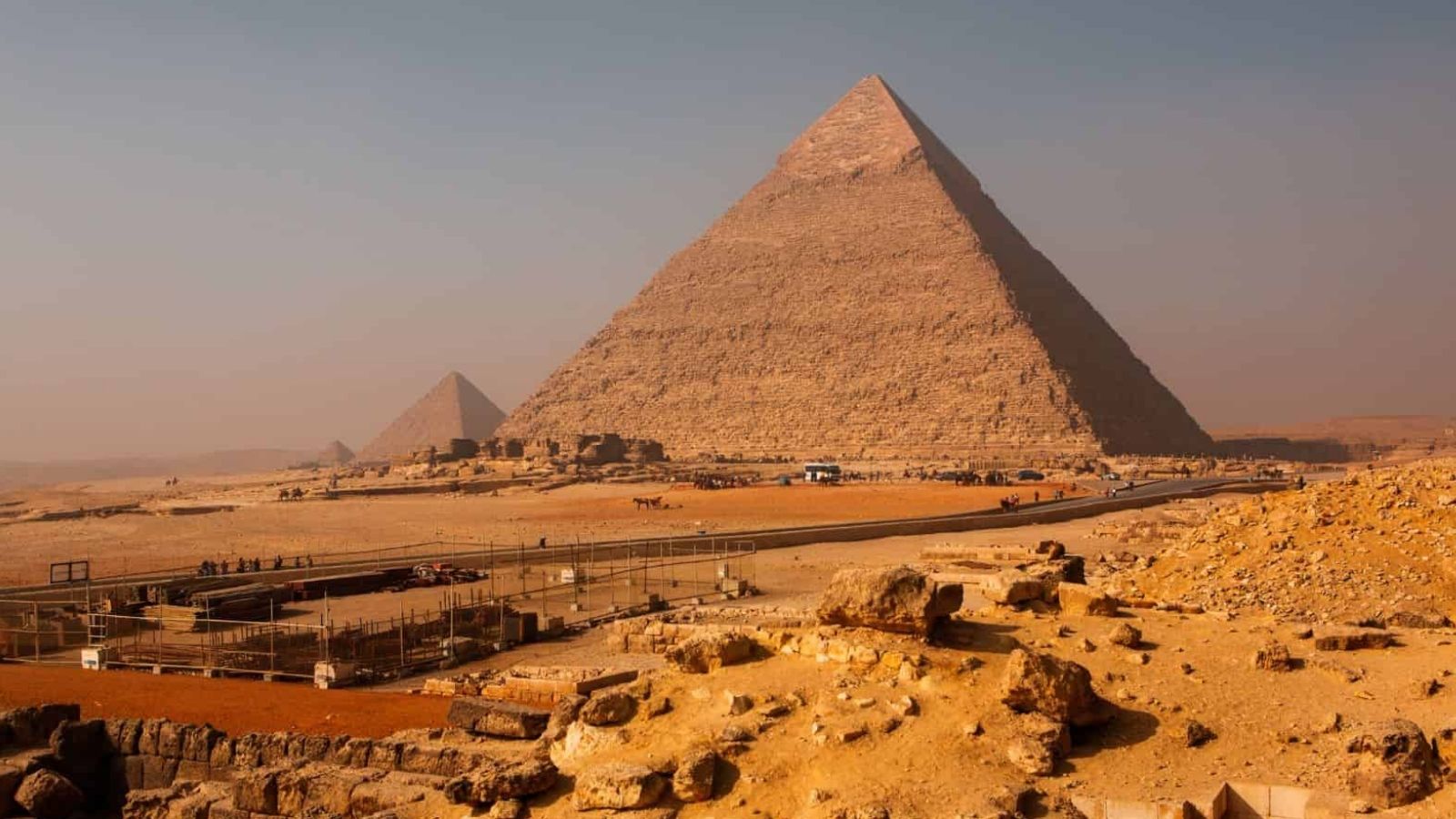 The Pyramids of Giza rise from a desert landscape beneath a hazy sky, with rocks and a fenced path in the foreground.