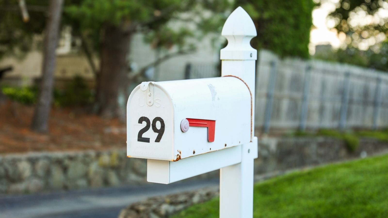 A white mailbox with the number 29 and red flag sits on a white post by a driveway next to a fenced yard.