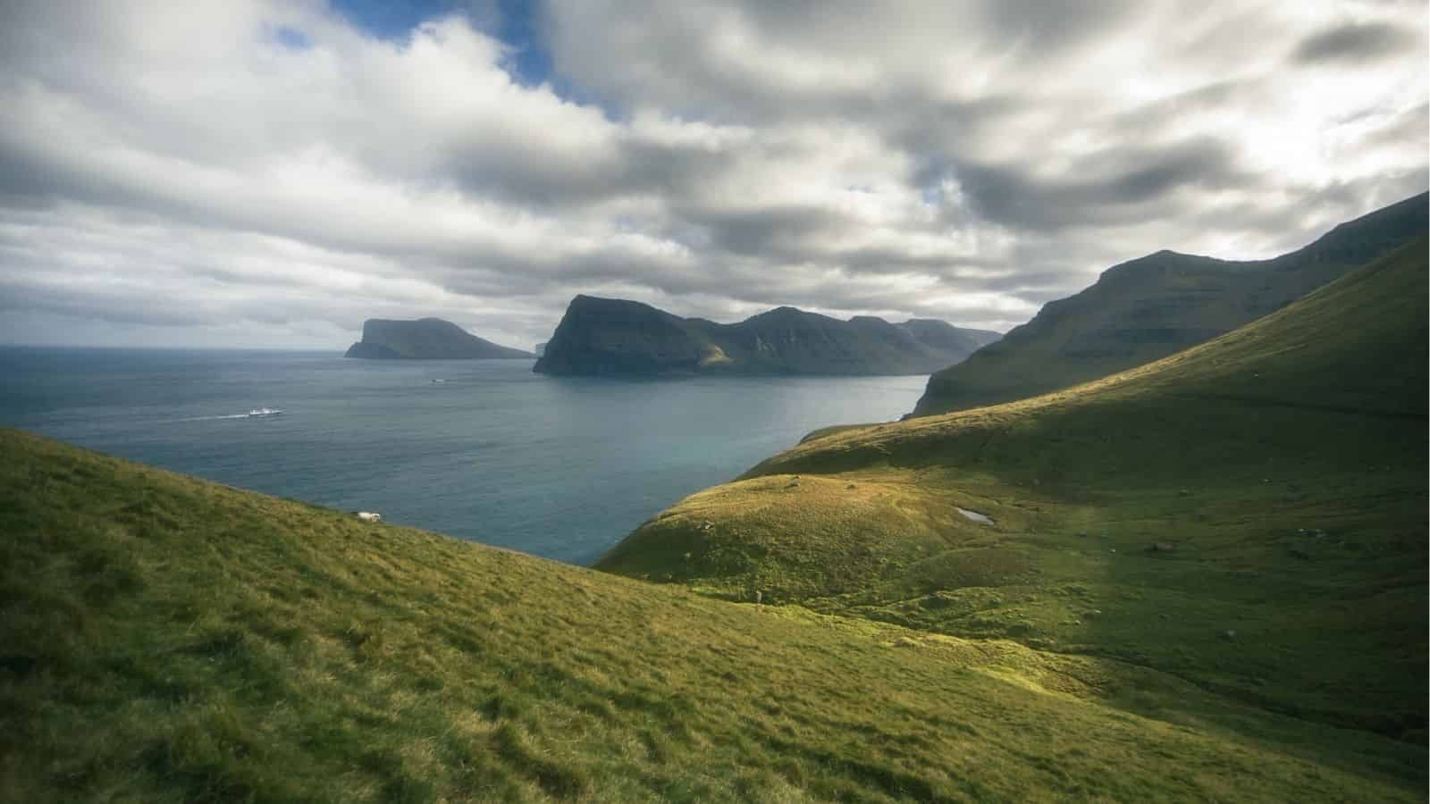 A grassy hill slopes to a calm sea with rocky islands and distant mountains beneath a cloudy sky.