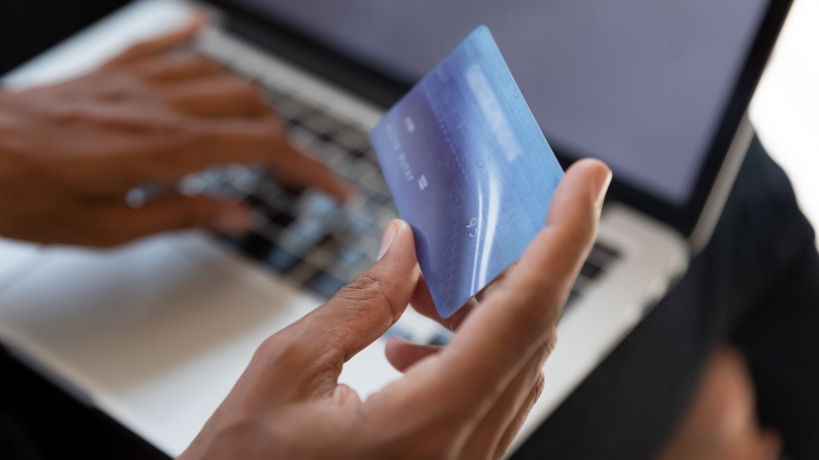 Someone types on a laptop while holding a blue credit card in their other hand.