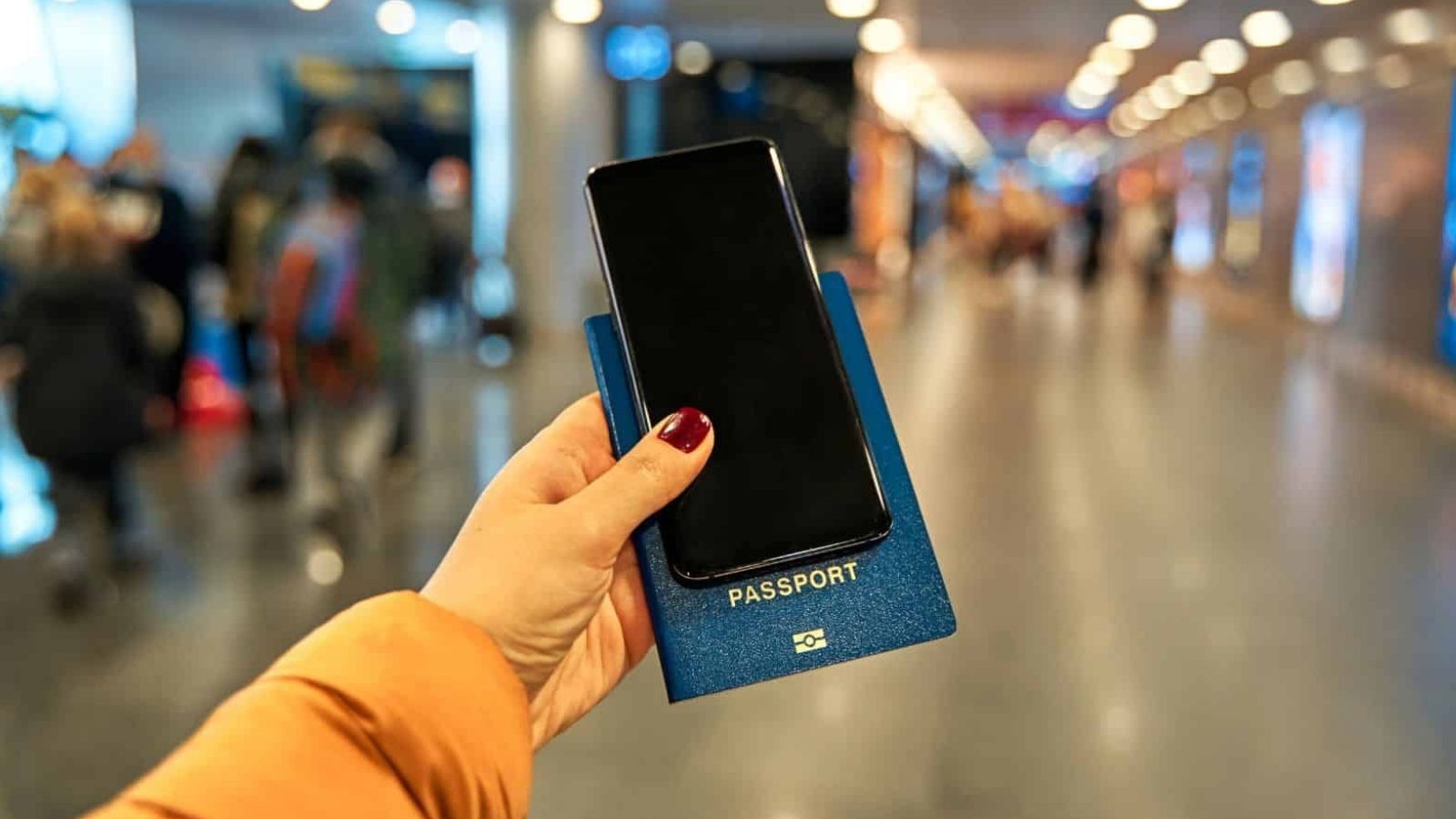 A person holds a smartphone and blue passport in an airport terminal with blurred lights and people behind them.