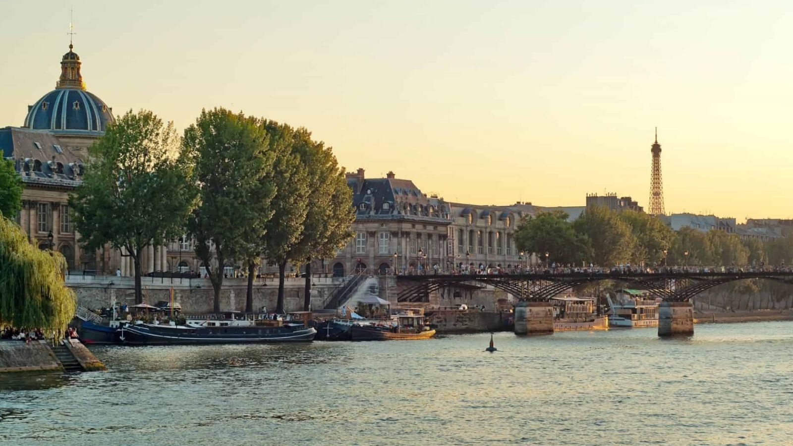 The Seine River in Paris at sunset, with docked boats, a tree-lined promenade, historic buildings, and the Eiffel Tower behind.