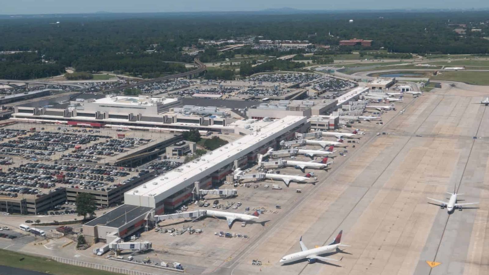 Aerial view of a busy airport terminal with several airplanes at gates and large parking lots behind.