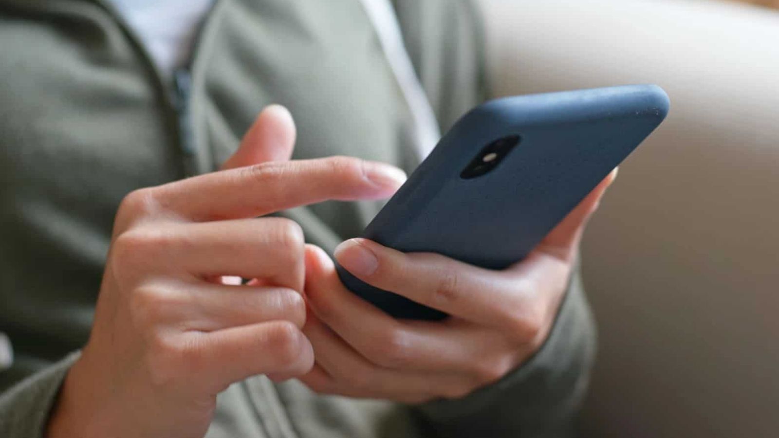 A person in a green jacket uses their right hand to operate a blue-cased smartphone.