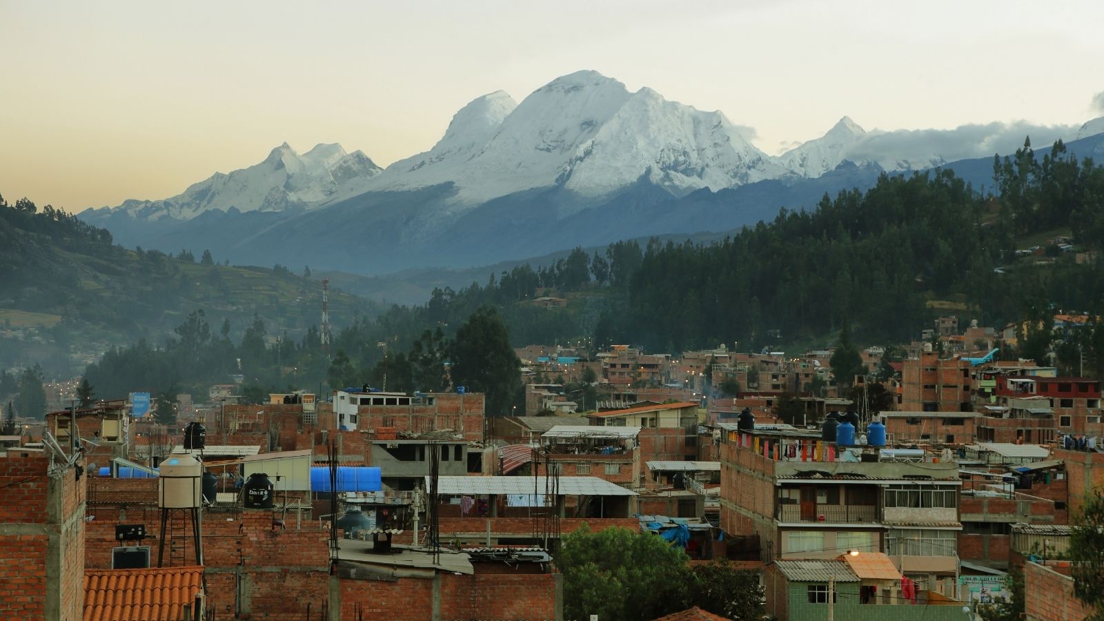 Brick buildings, trees, hills, and snow-capped mountains sit under a cloudy sky in this layered city landscape.