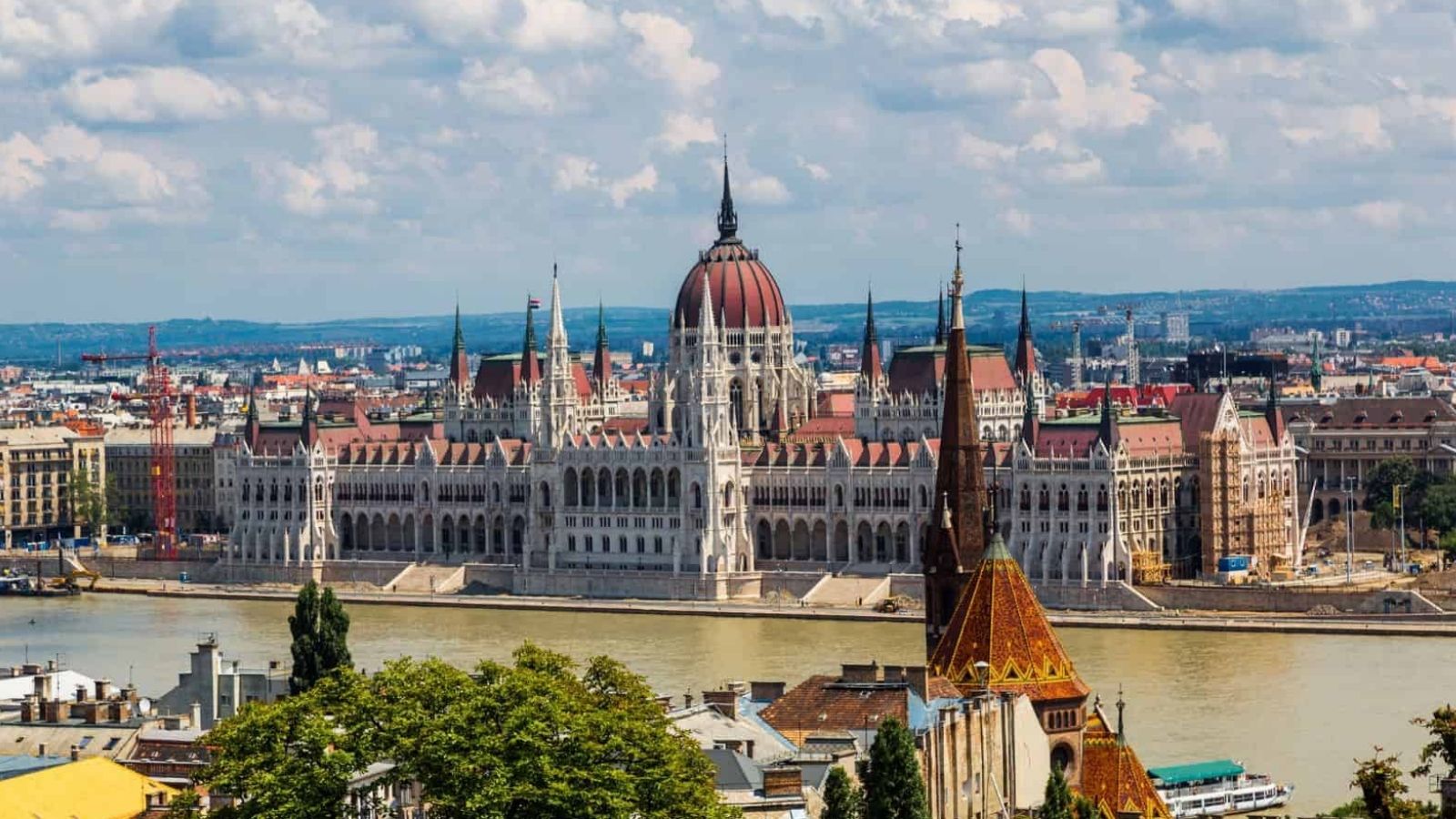 The Hungarian Parliament Building is next to the Danube River in Budapest, framed by cityscape and clouds.