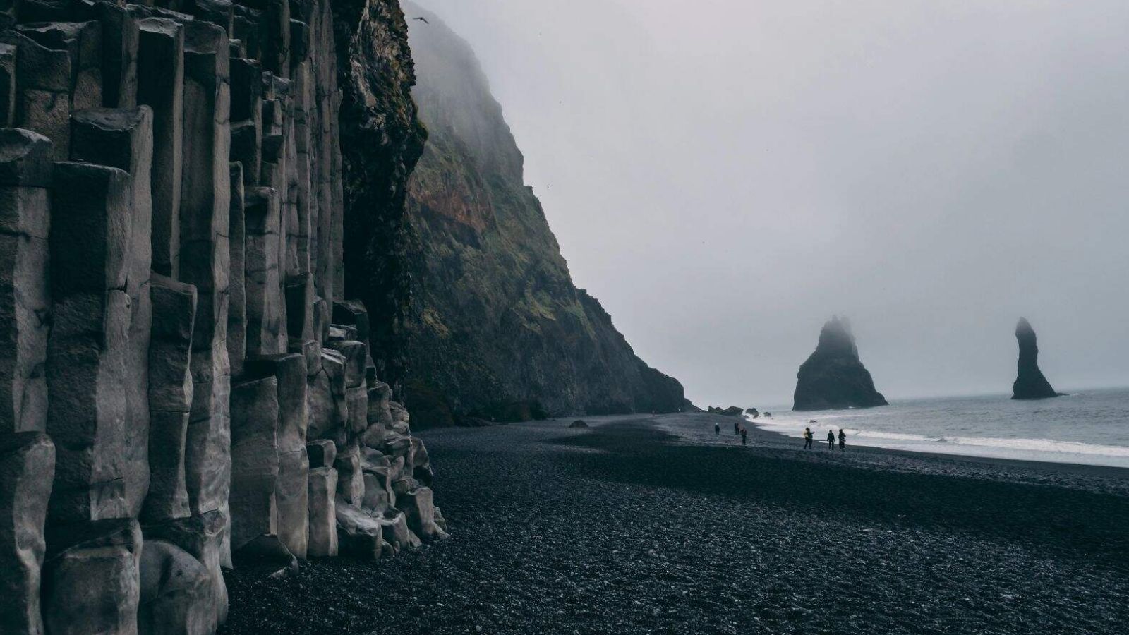 Mist shrouds a black sand beach, basalt columns line the left, sea stacks rise in the distance, and people walk the shore.