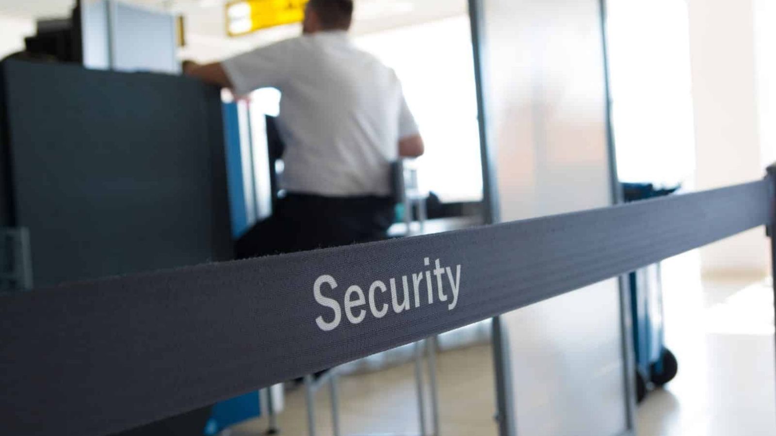 A black "Security"-labeled barrier stands before a blurred checkpoint and a uniformed person in the background.