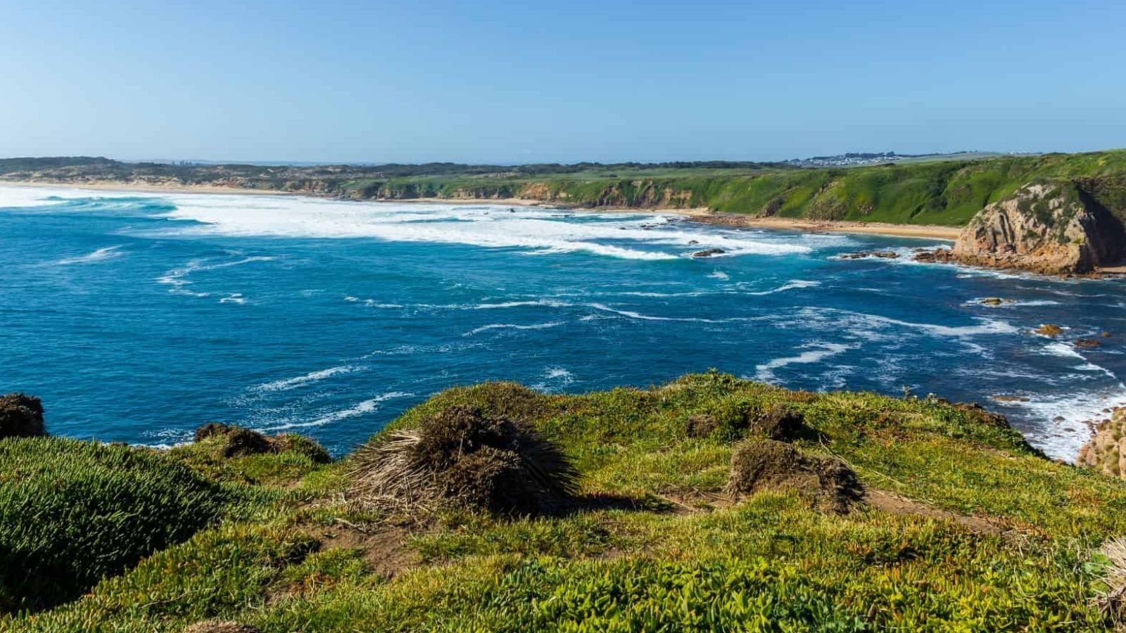 Blue ocean waves crash against rocky cliffs and green hills beneath a clear blue sky.