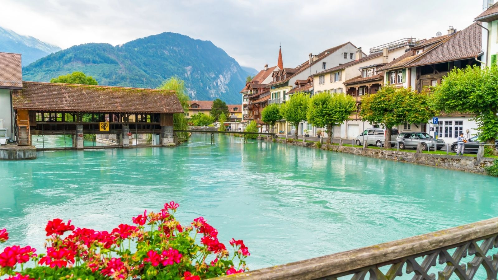 A turquoise river flows by traditional buildings and a wooden bridge, with red flowers in front and tree-covered mountains behind.